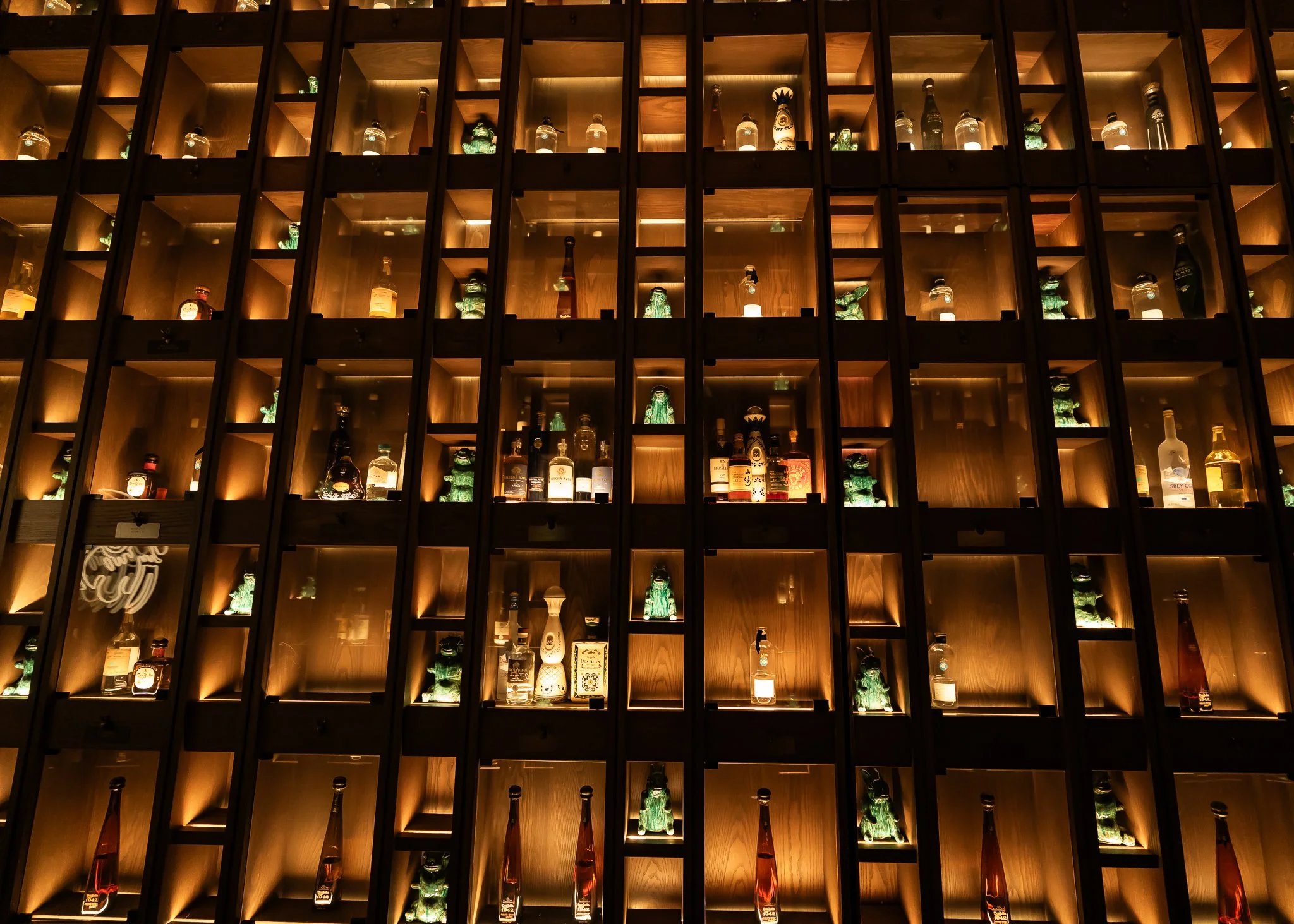 A wall with multiple wooden compartments displaying various liquor bottles and decorative ceramic figures, illuminated by warm lighting.