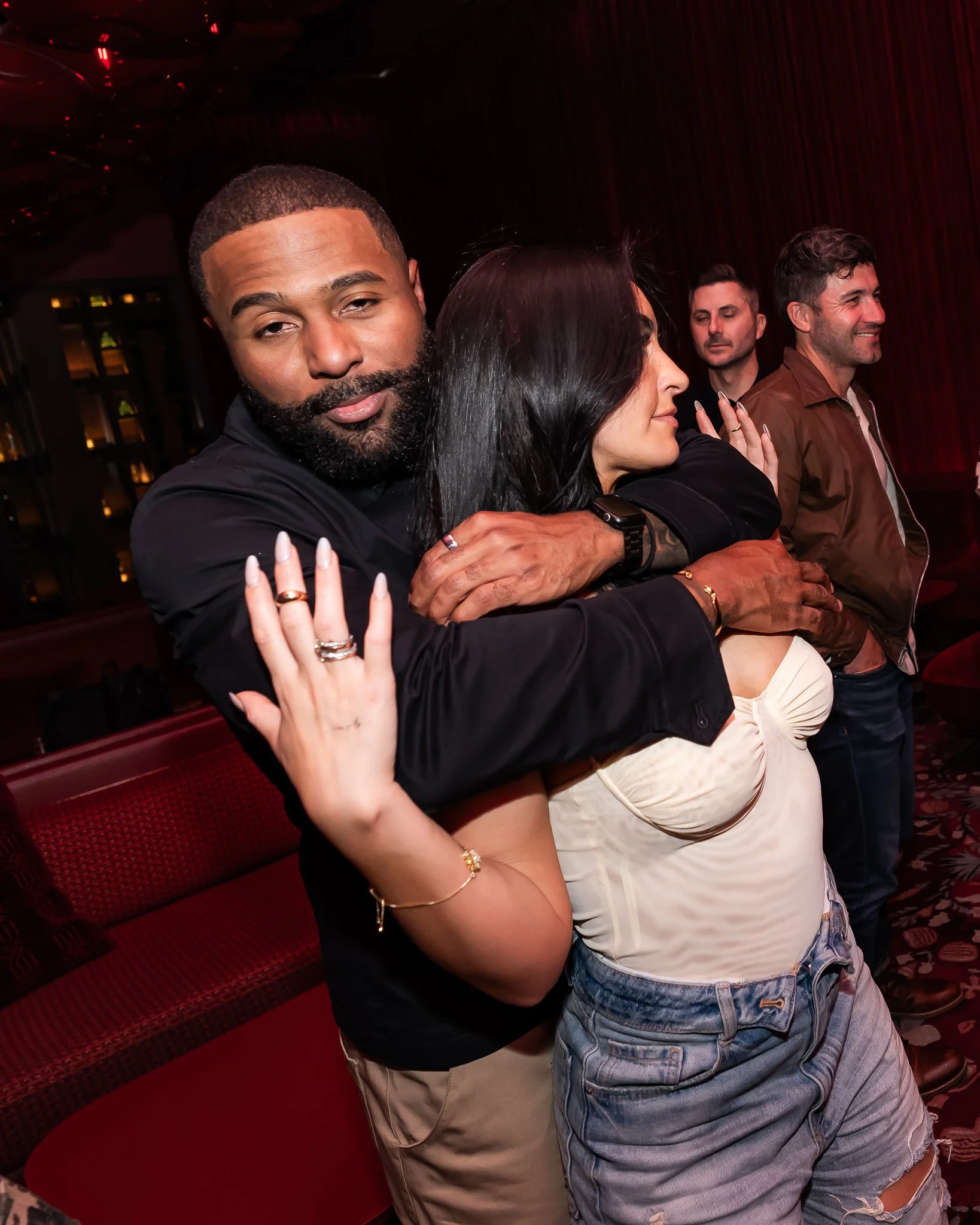 A group of five people are standing close together in a dimly lit room with dark red curtains and a patterned carpet. The focus is on a man with short hair and a beard hugging a woman with long dark hair, both smiling softly. The people behind them a