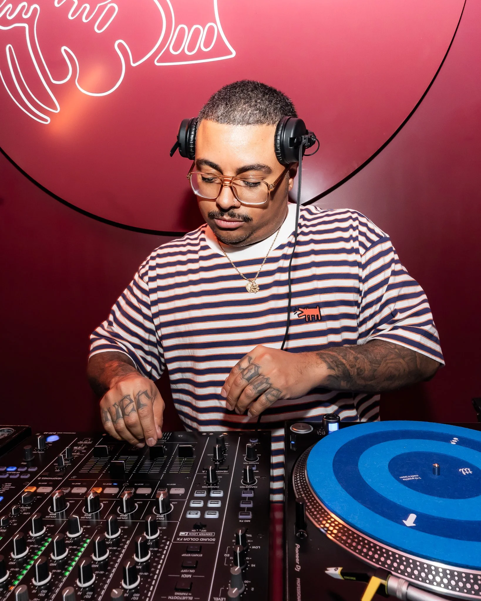 A man with short hair, glasses, and tattoos on his hands is DJing using a mixer and turntable. He is wearing a striped shirt and headphones, with a pink background and neon artwork behind him.