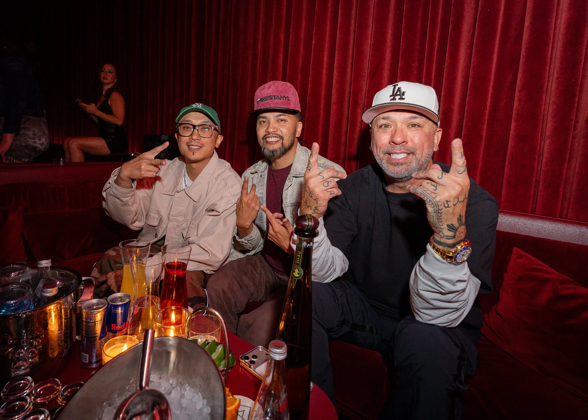 Three men sitting at a table with colorful drinks in front of them, smiling and making gestures, in front of red velvet curtains. A woman in black and gray attire is sitting in the background.