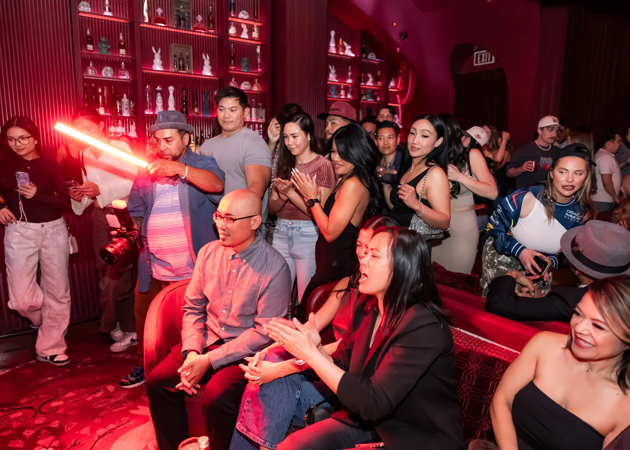 A diverse group of people at a social gathering or party in a dimly-lit, red ambiance venue. Some are seated, others standing, and some are looking at their phones or engaging with each other. The background shows a red wall with shelves of bottles a