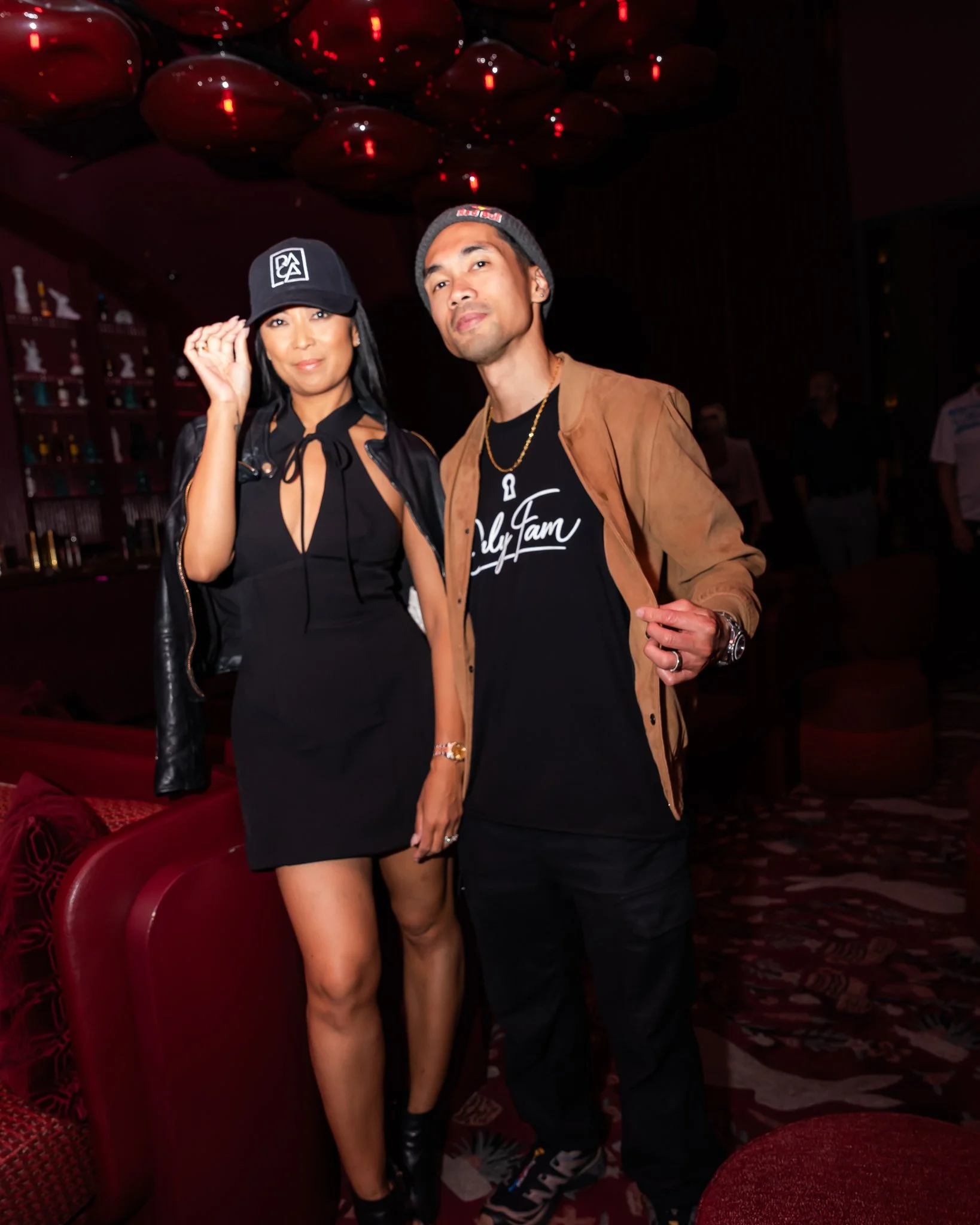 A man and woman posing together indoors at night, with red lighting and decorative elements in the background. The woman is wearing a black dress, a black cap, and black thigh-high boots, while the man is wearing a tan jacket, black t-shirt, and a be