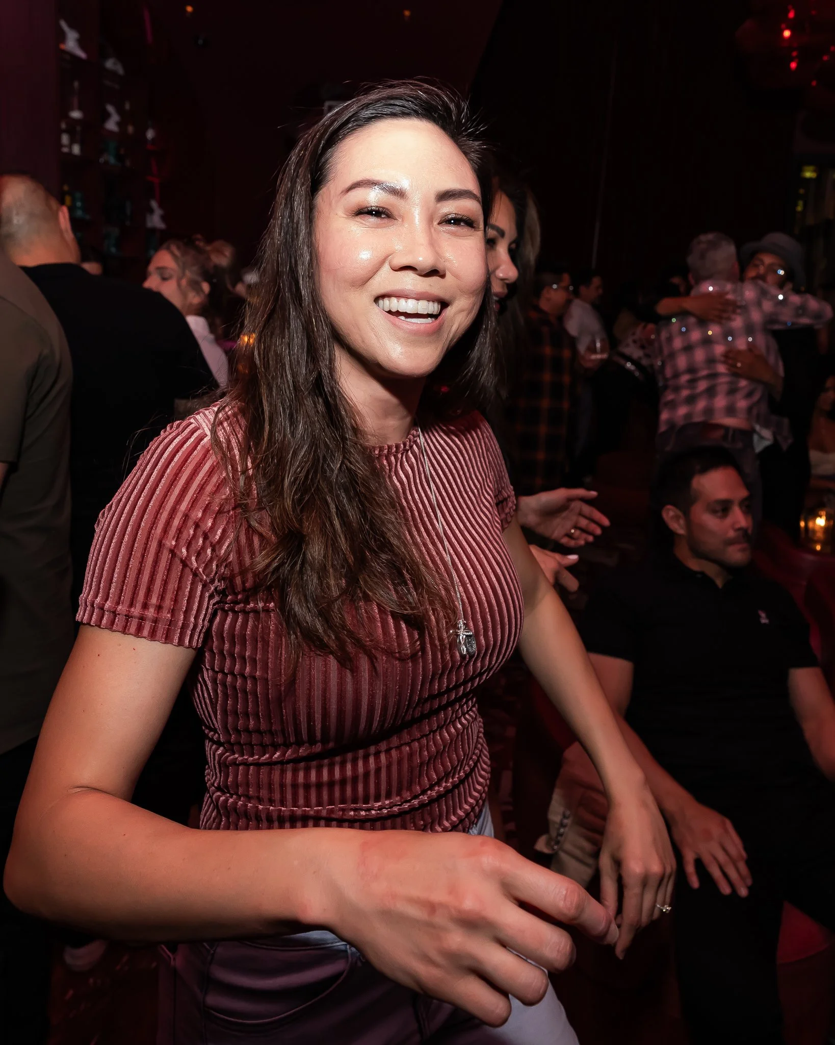 A woman smiling at the camera with people hugging in the background at a social gathering.