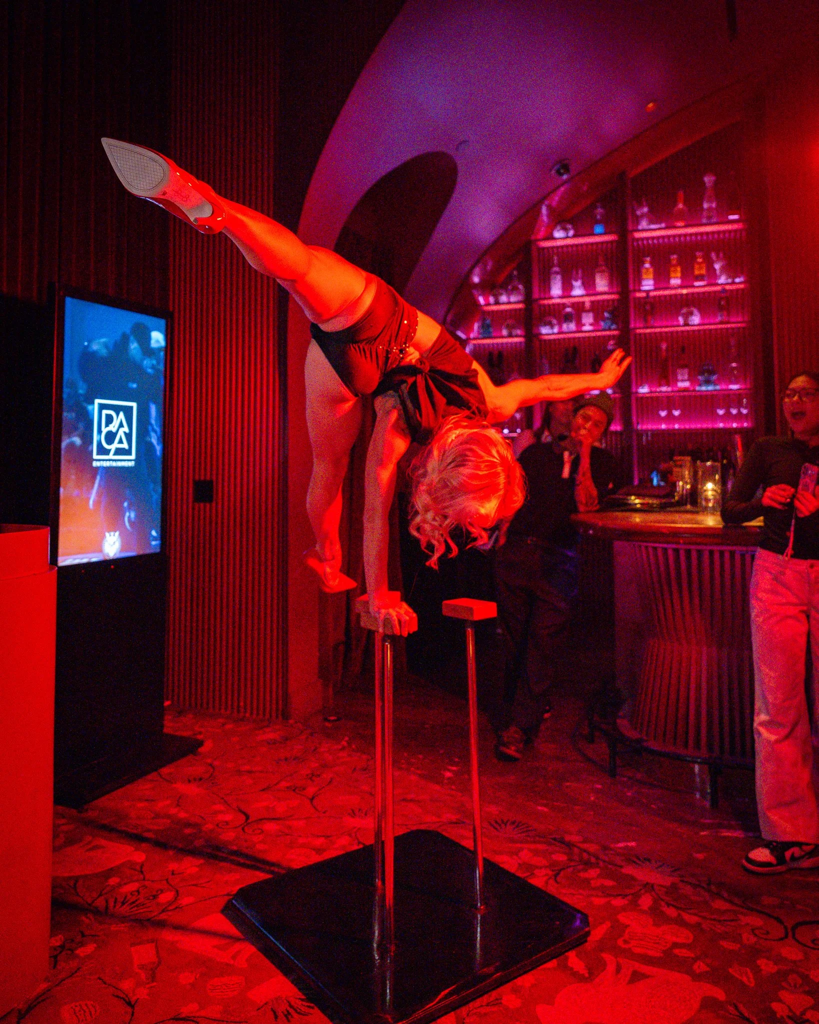 A performer is balanced on their hands on a raised platform, with their legs extended vertically and resting on a pole, in a dimly lit, red-hued venue with a bar and an audience watching.