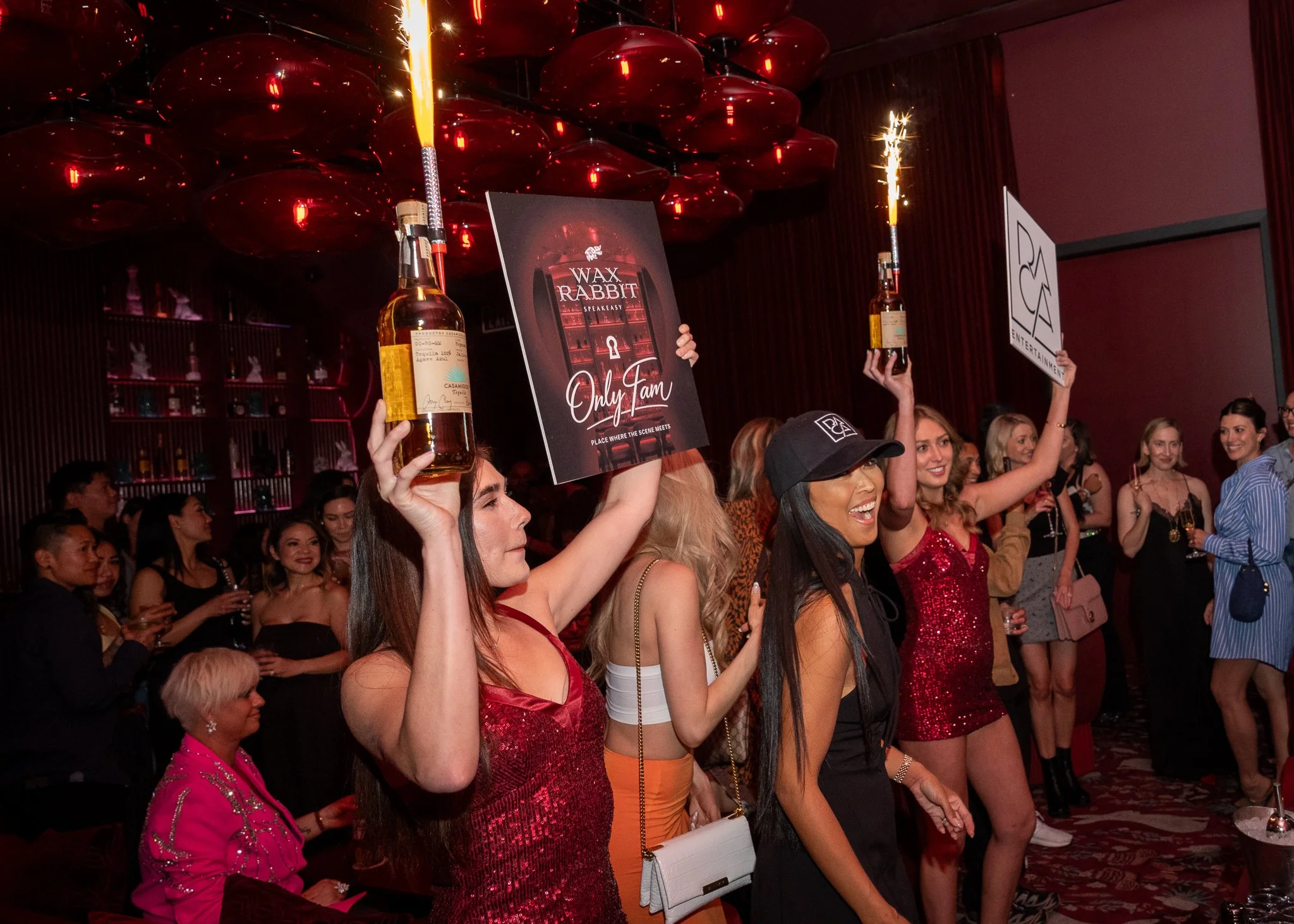 A group of women at a celebration event holding bottles of alcohol and signs, with sparklers and a dark red and black themed background.