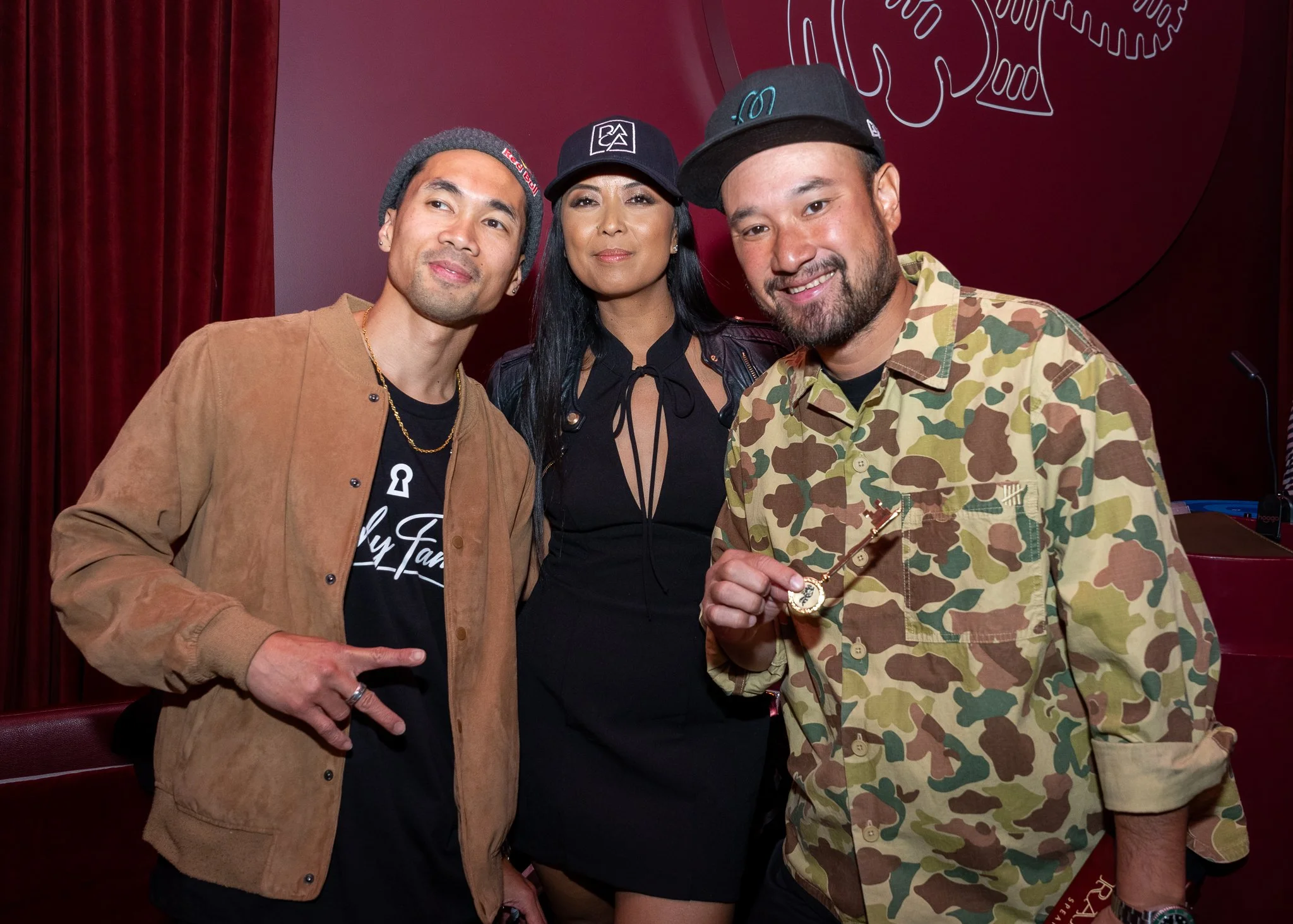 Three people posing together at an indoor event, with red curtains and a decorative line drawing on the wall behind them. The person on the left is a man wearing a gray beanie, tan jacket, and black shirt, pointing at his waist. The person in the mid