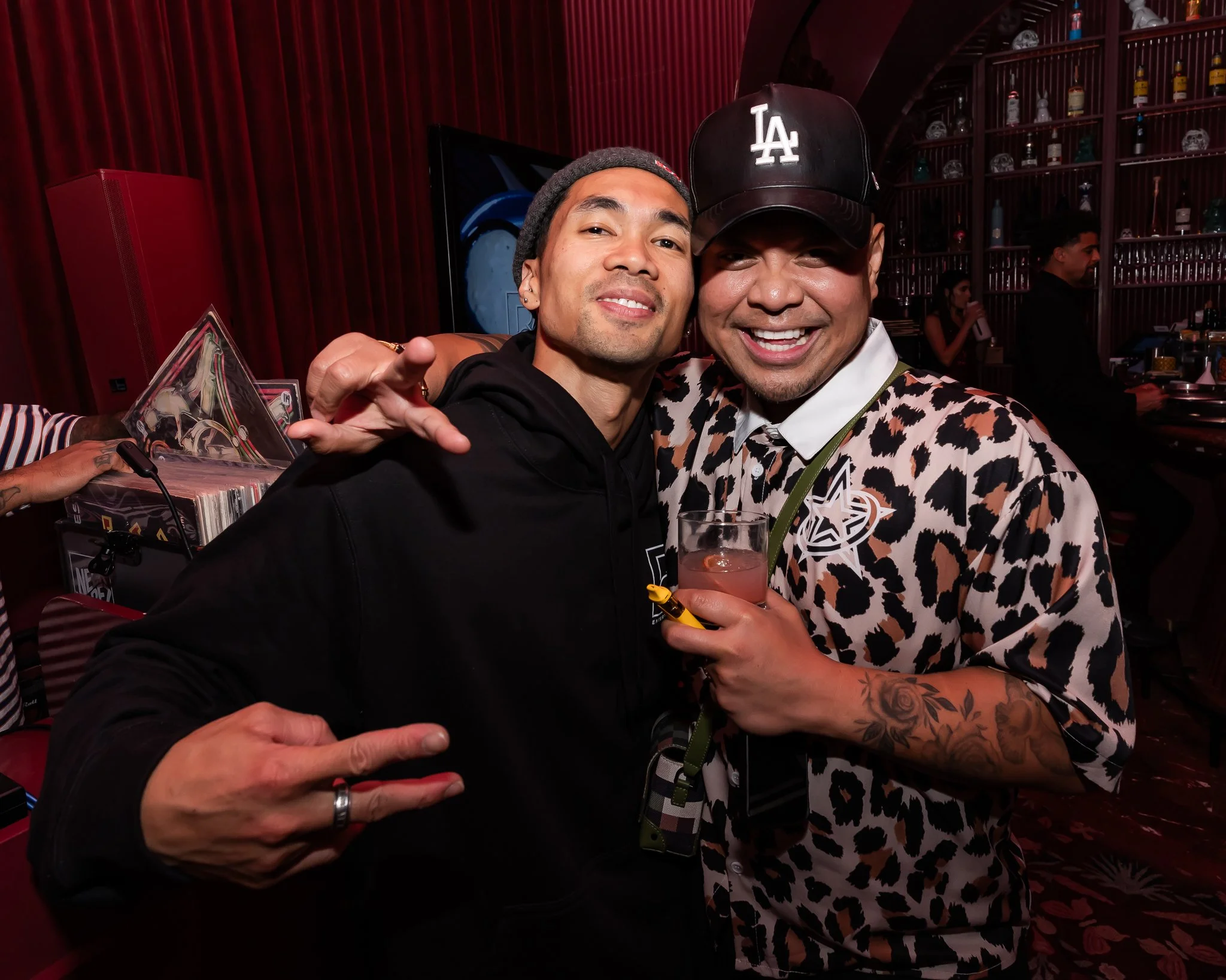 Two men smiling and posing closely together at a social event or party, with one holding a drink and the background showing a bar with bottles and skull decorations.