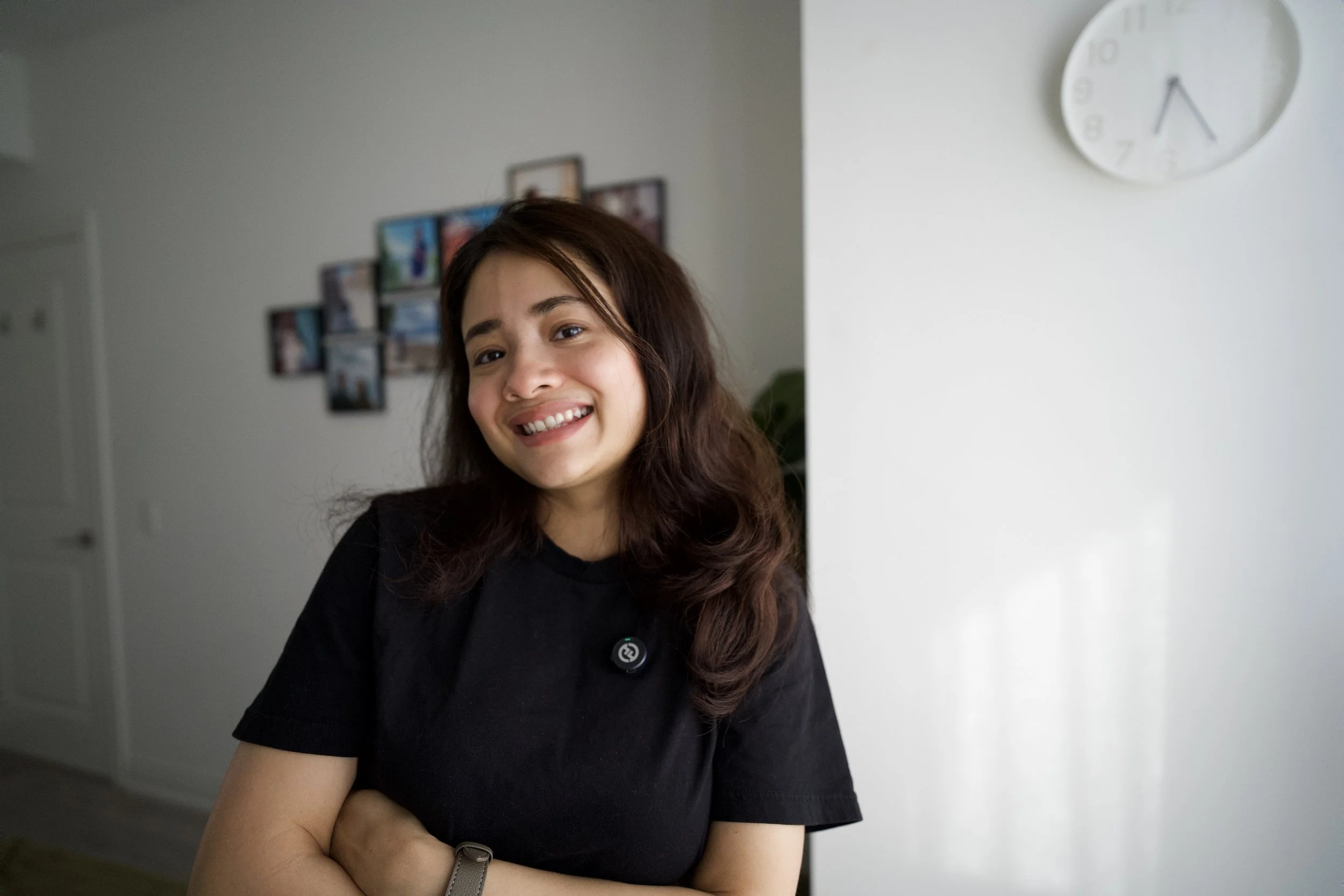 A smiling woman with long brown hair wearing a black shirt standing in a room with white walls, a picture collage on the wall, and a round white clock showing 3:25.