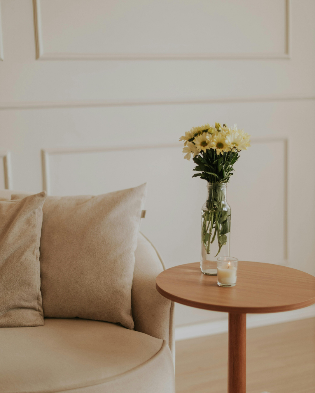A living room side table with a vase of white and yellow flowers and a lit candle, next to a beige sofa with matching cushions, in a minimalist decor setting.