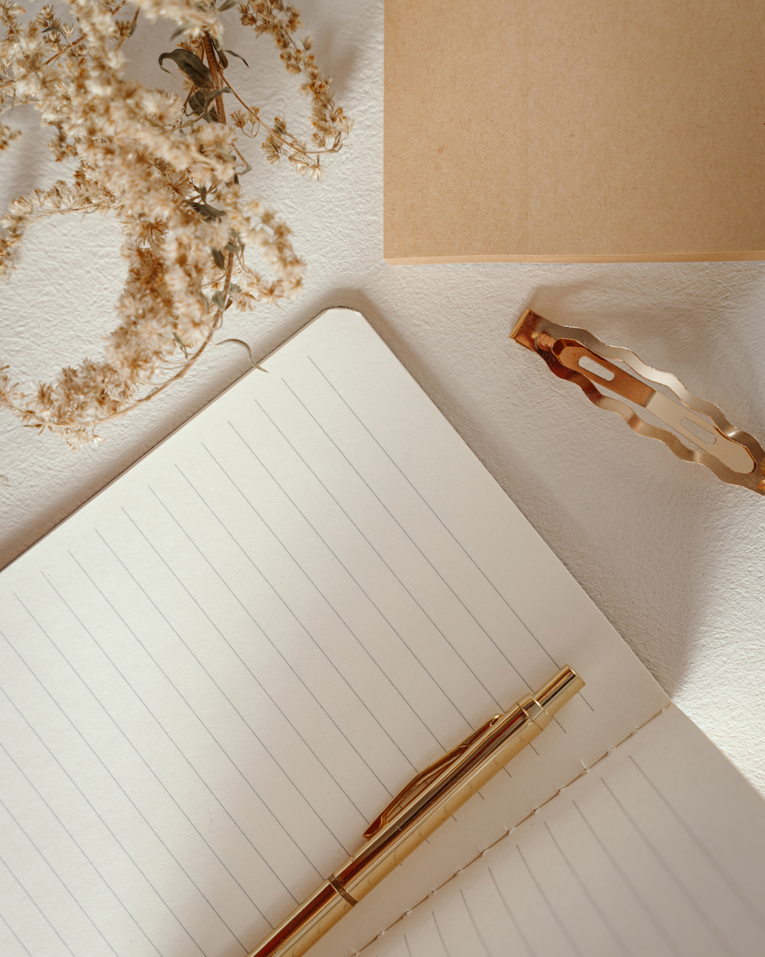 Open notebook with lined pages and a gold pen, dried flowers, a gold hair clip, and a brown paper sheet on a white textured surface.