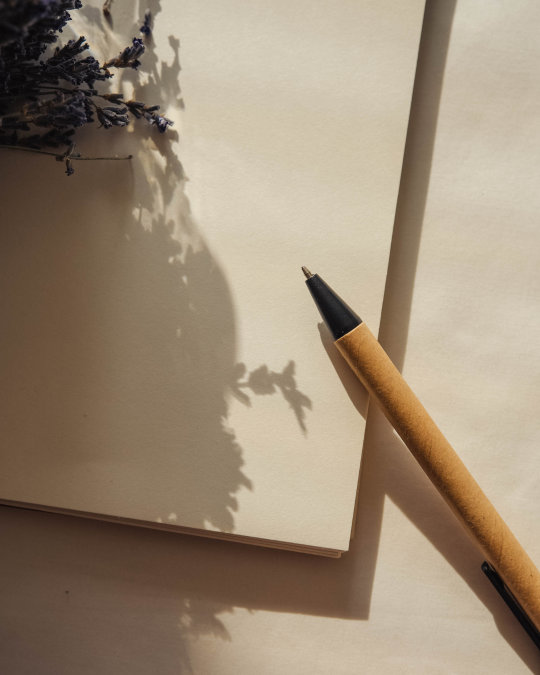 A blank notebook with shadows of lavender attached to its top left corner, a black and brown pen resting on the right side of the notebook, sunlight casting shadows on the notebook and the surface below.