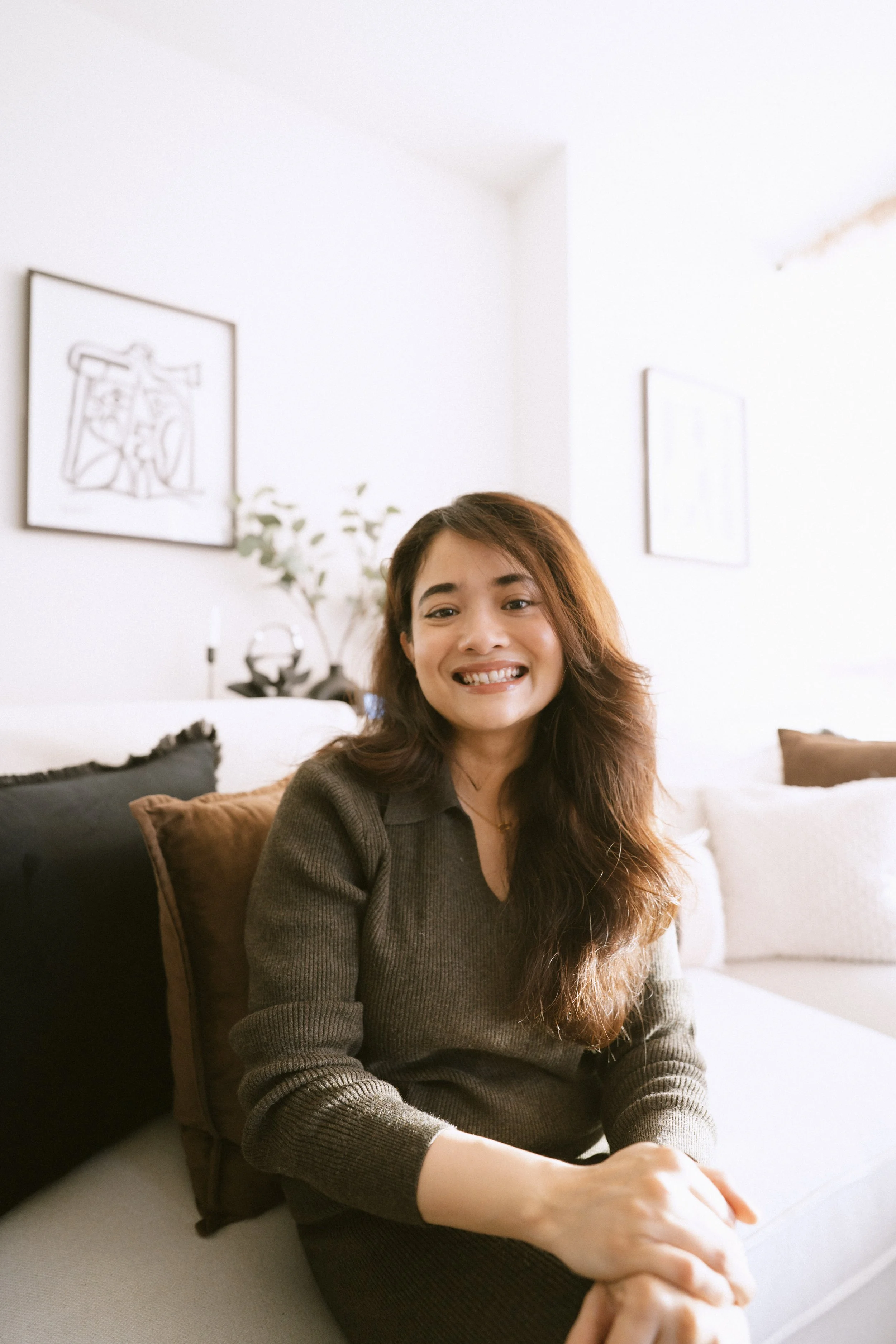 A woman with long brown hair smiling while sitting on a couch with brown and black pillows in a bright living room.