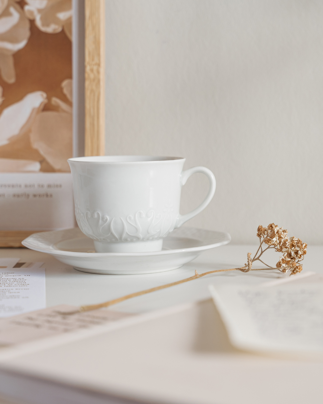 White ceramic teacup on a saucer with a dried flower and papers nearby.