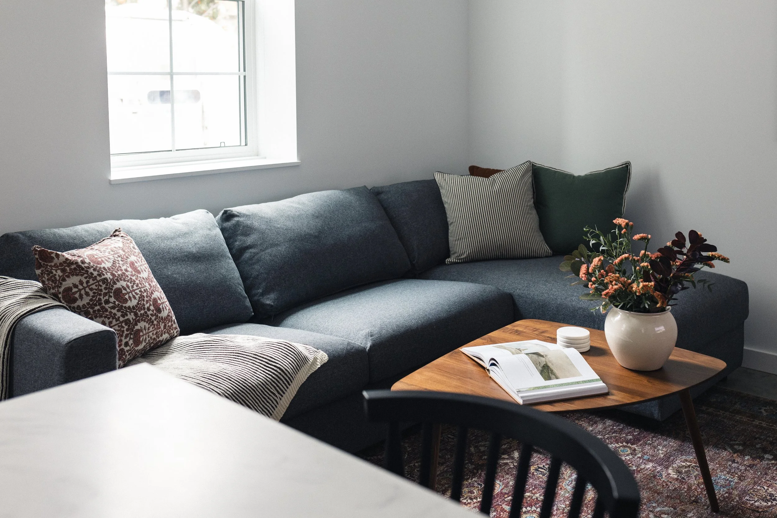 A cozy living room with a blue sectional sofa bed, decorative pillows, large screen smart TV, reading material, and copious amounts of natural light.