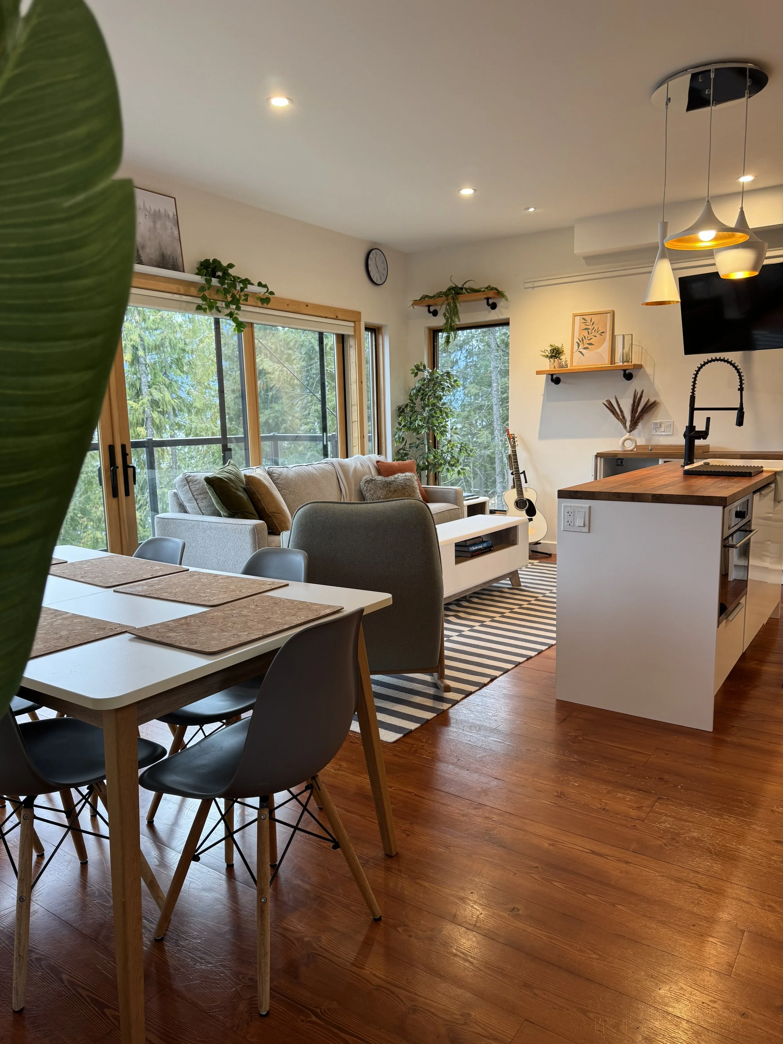 A cozy, modern living room with large windows overlooking trees. It includes a dining table with chairs, a sofa with cushions, a striped rug, a guitar, and decorative shelves with plants and artwork. The space has wooden flooring and contemporary lig