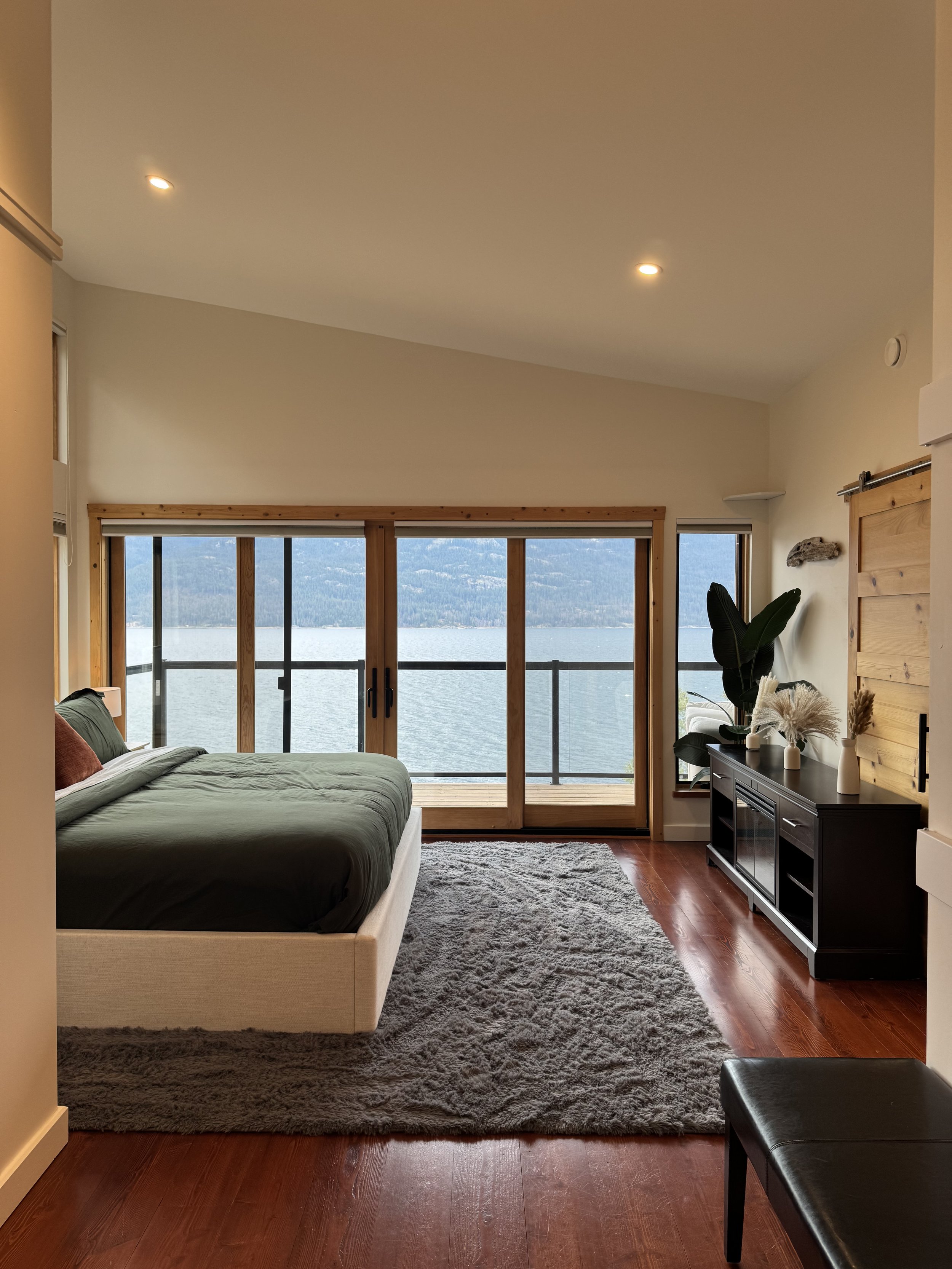 Modern bedroom with large sliding glass door overlooking a lake, featuring a King Size bed with green bedding, a gray rug, a fireplace with decorative vases, and indoor plants.