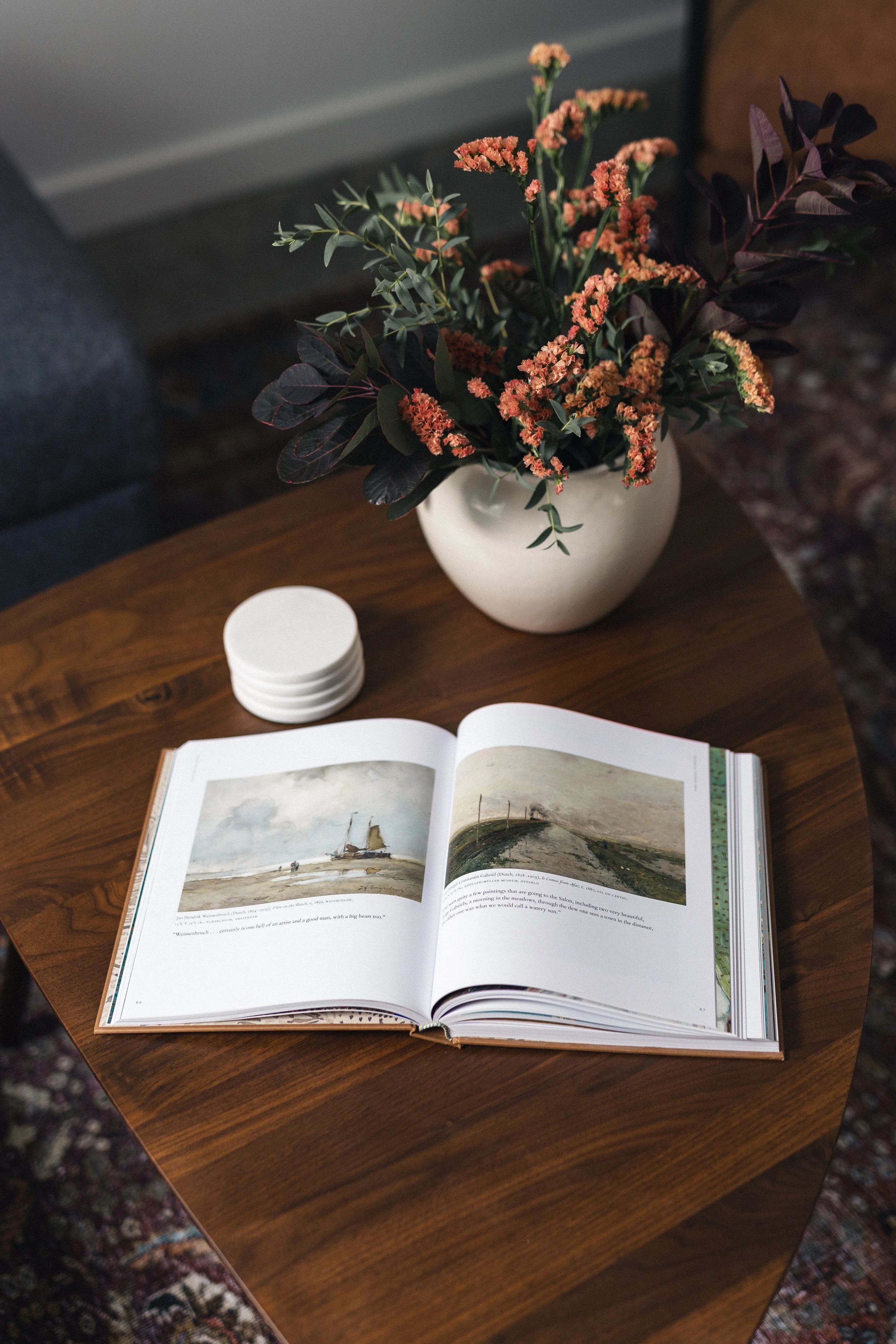 Open art book on a wooden table with a white vase containing pink and green floral arrangement and a stack of white coasters.