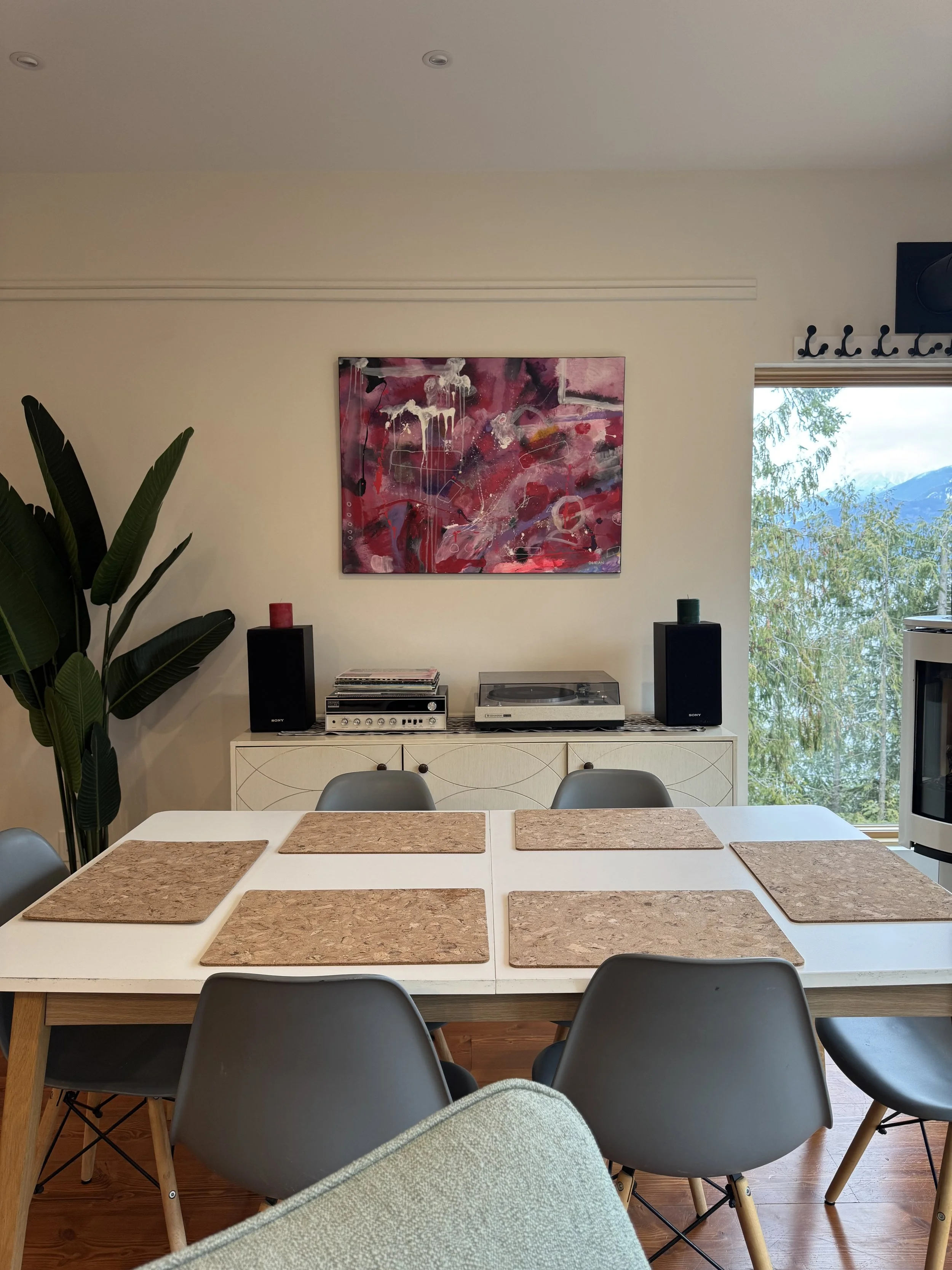 A dining area with a white table set with six brown placemats, surrounded by six gray chairs. On the back wall, there is a cabinet with a turntable, stereo, and speakers. A large abstract painting with pink, red, and white colors hangs above the cabi