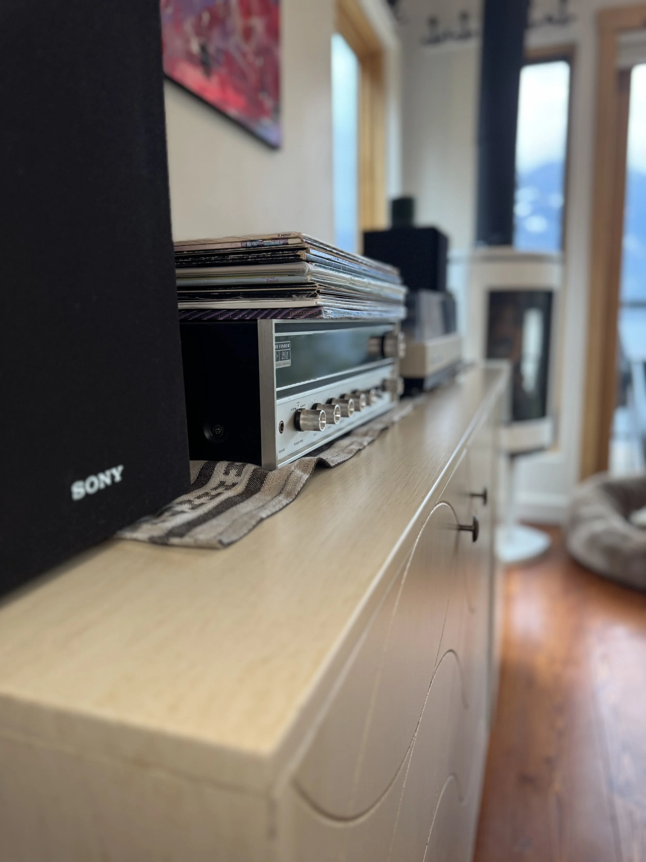 A close-up of a vintage stereo receiver with a stack of vinyl records on top, on a light wooden sideboard in a living room.