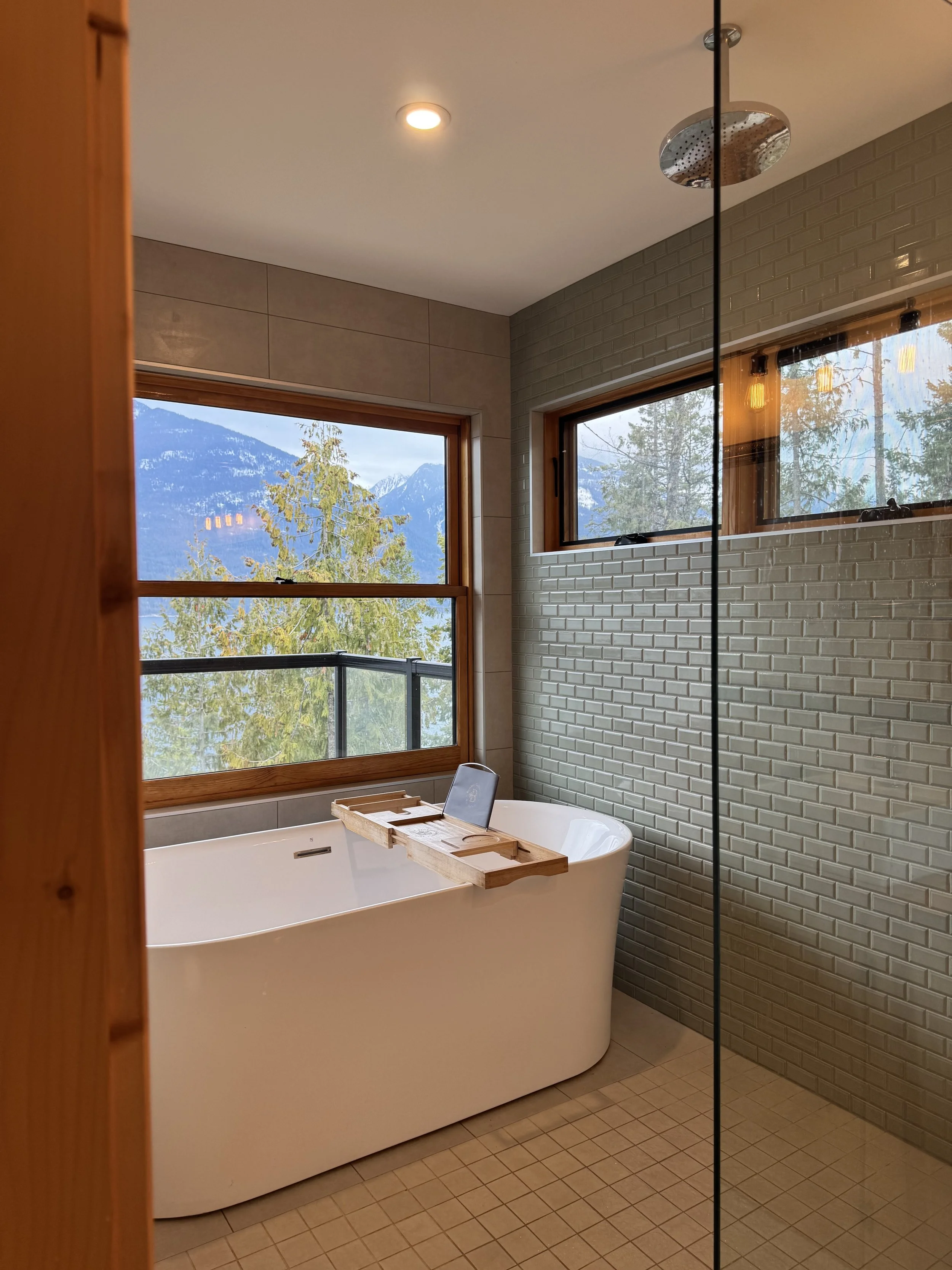 Modern bathroom with a large bathtub positioned in front of a window with mountain and tree views. The bathroom has beige tiles and textured gray tiles on the walls, with a ceiling-mounted shower head and a small window on the right.