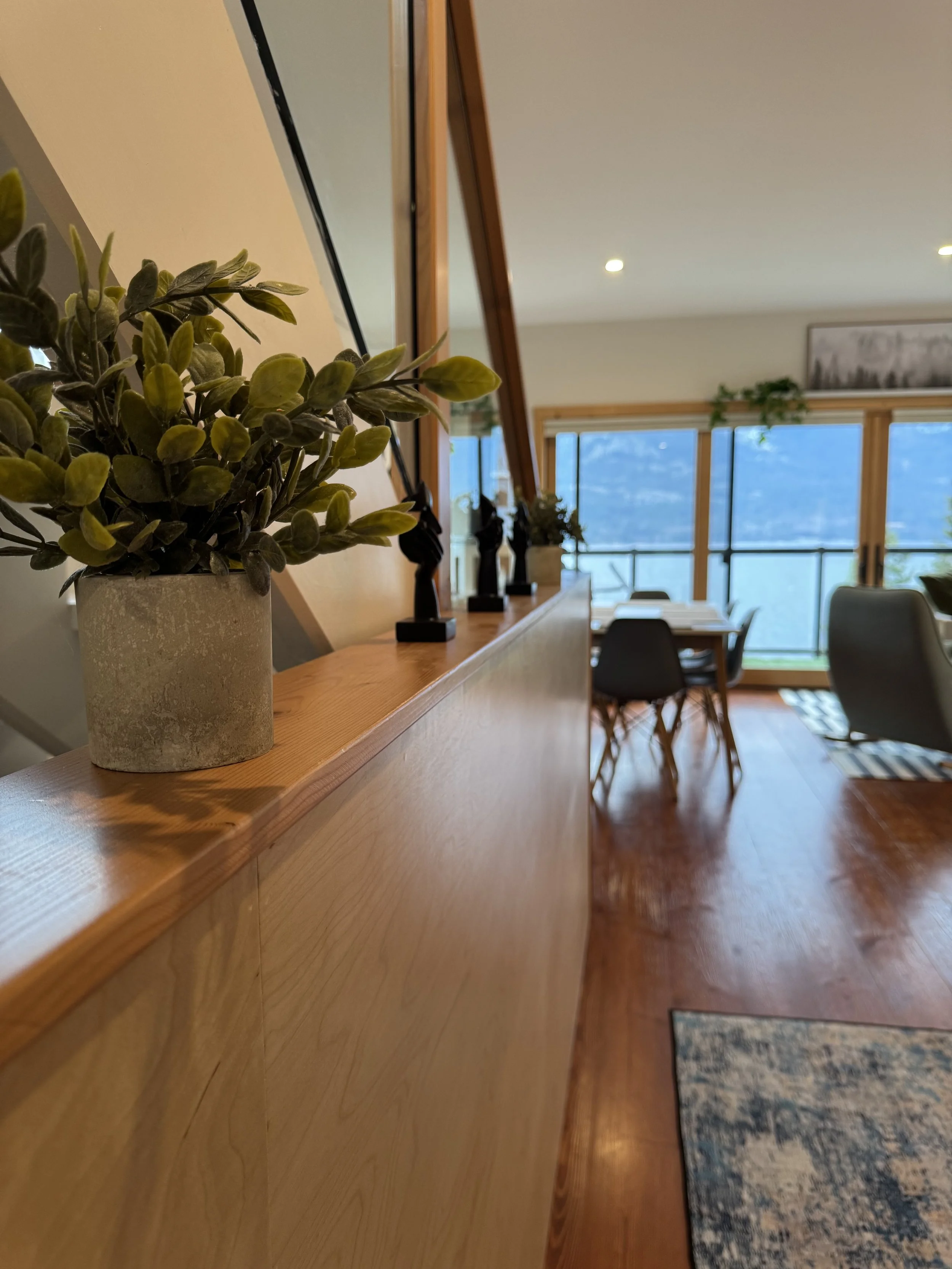 Interior of a modern living and dining area with wooden floors, a wooden partition, potted plants, and tables and chairs near large windows.