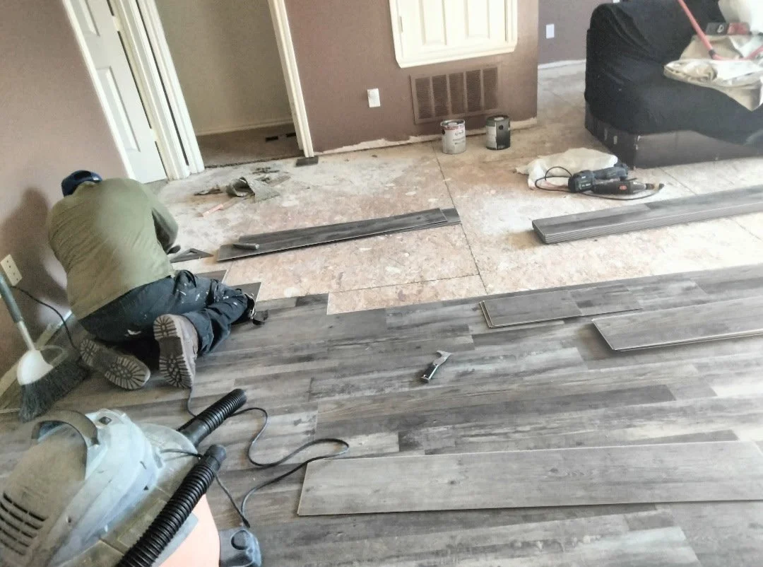 A person installing new flooring in a room, with wooden planks, tools, and supplies scattered around.