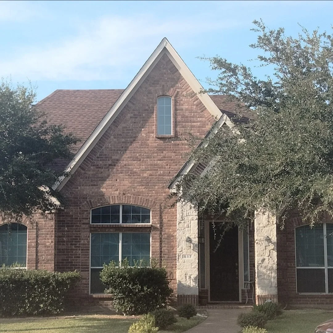 A brick house with a steep roof, a tall arched window above the entrance, and bushes in front.