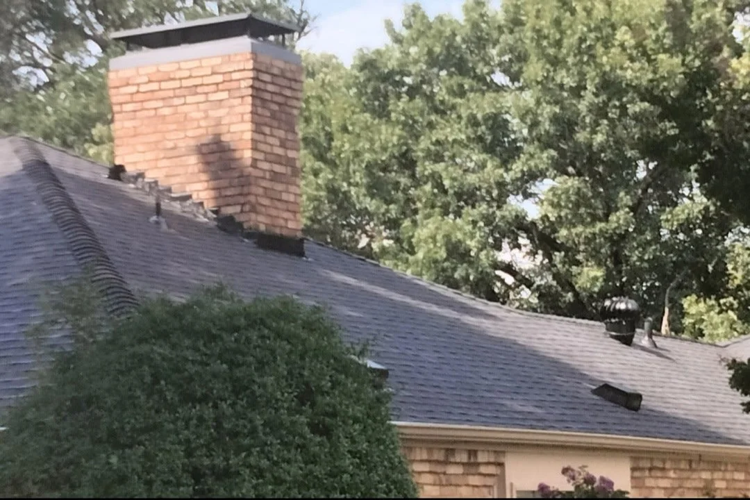 Close-up of a roof with missing shingles, a brick chimney, and trees in the background.