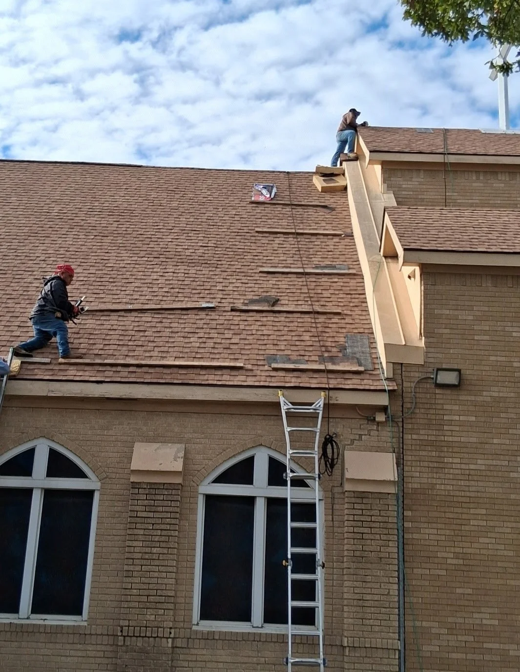 Workers repairing or installing a roof on a large brick building with windows, using safety equipment and ladders, under a partly cloudy sky.