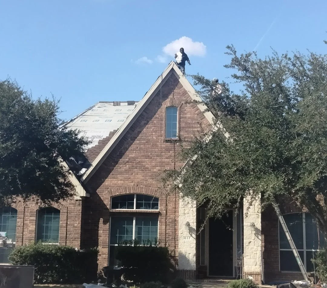 Construction workers on the roof of a brick house during roofing repair, with ladder leaning against the side and trees partially obscuring the view.
