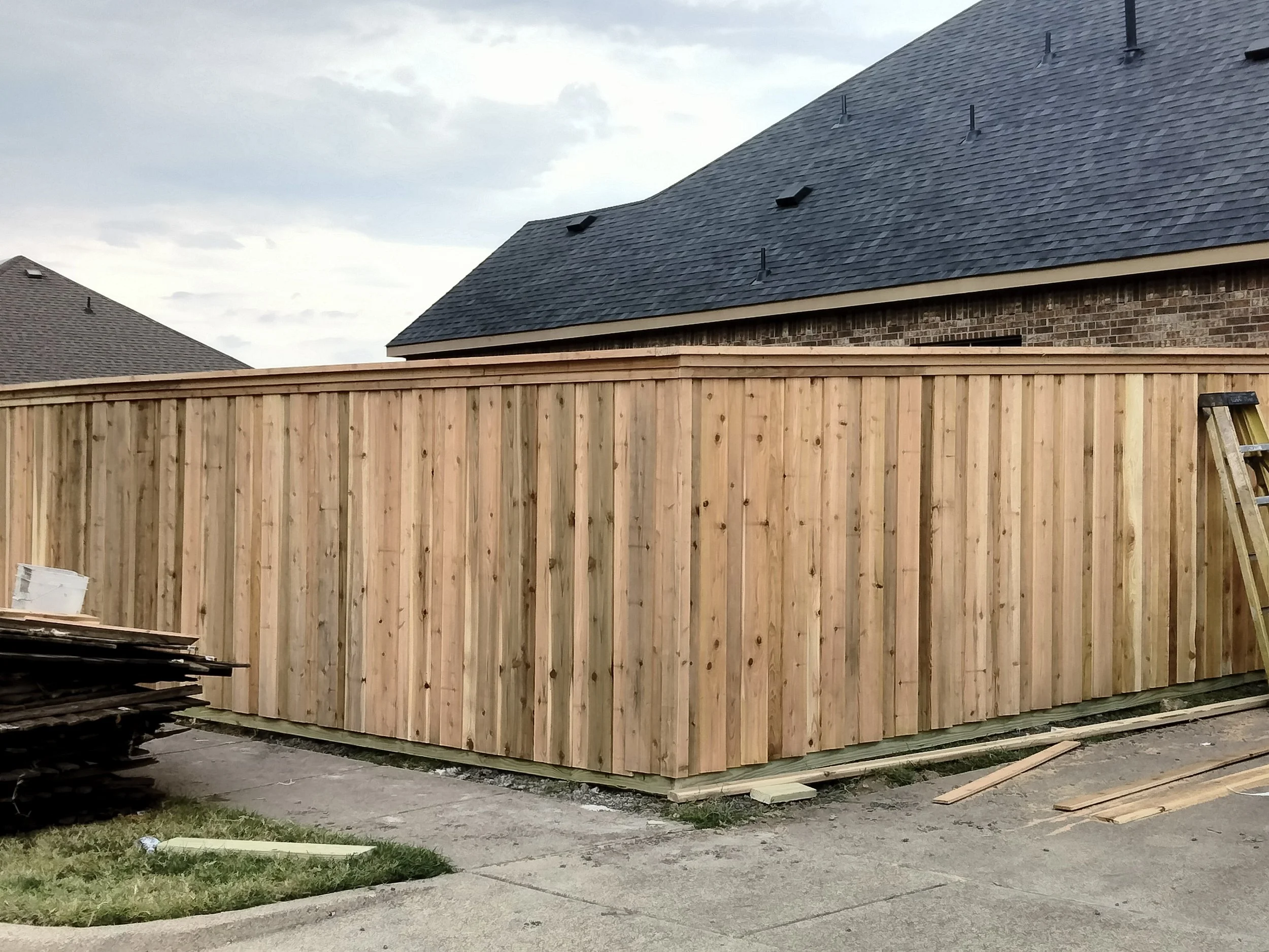 A wooden privacy fence under construction in a residential backyard, with a ladder leaning against it and building materials nearby, adjacent to brick houses with dark shingle roofs.