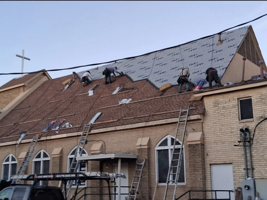Workers installing new roofing on a church building, with ladders leaning against it, and a cross visible in the background.