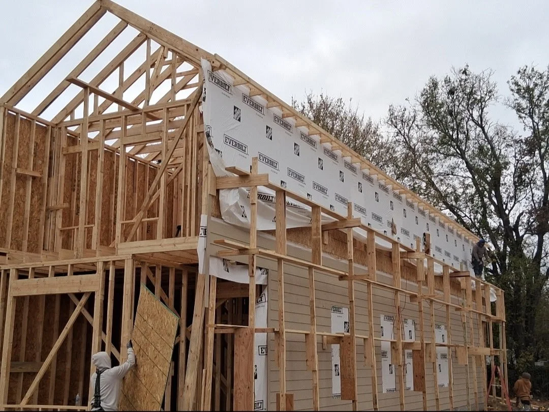A house under construction with a wooden frame and partial exterior wall panels. Two workers are seen working on the structure, with one on the ground installing sheathing and another on the upper level.