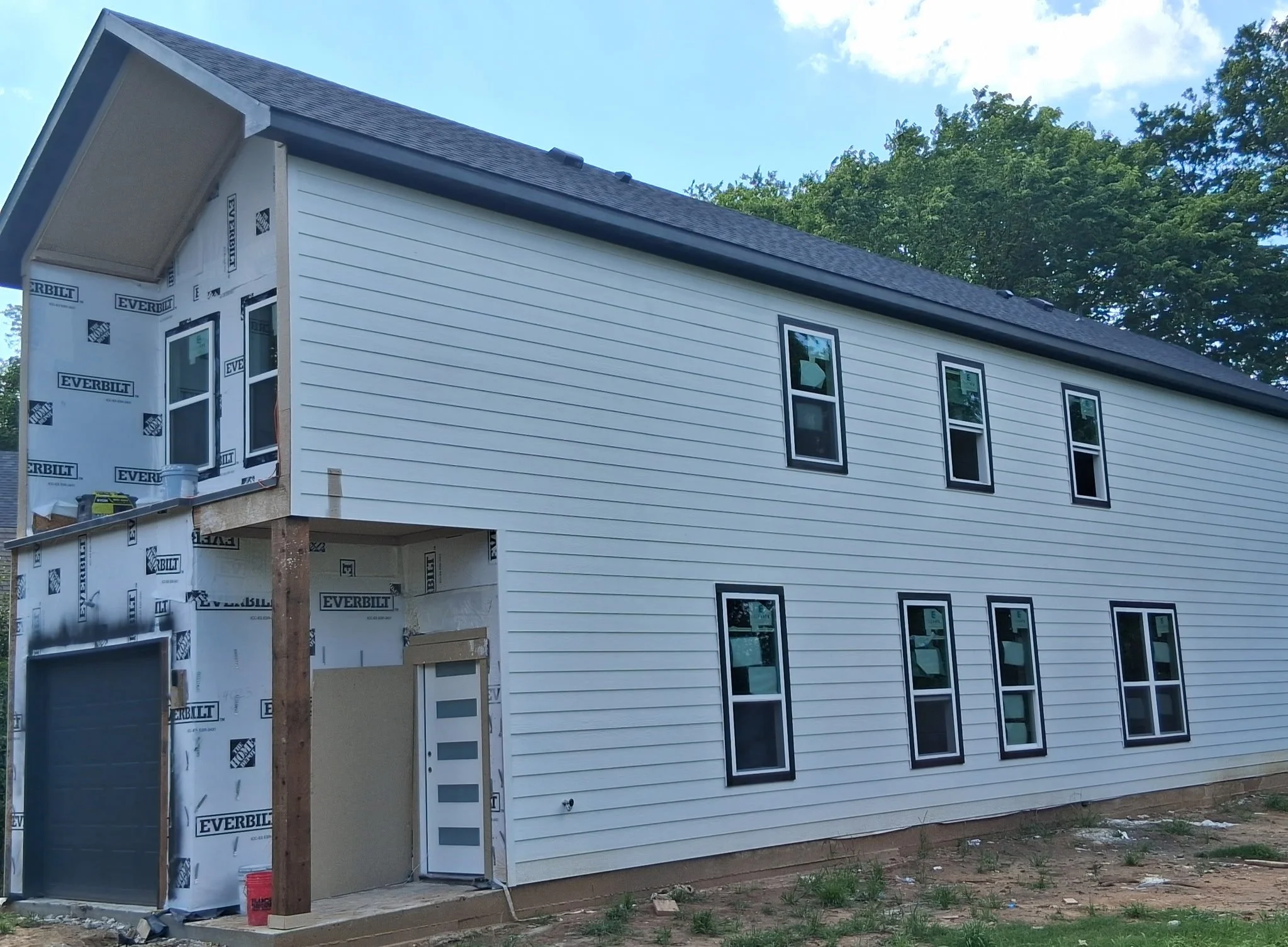Side view of a house under construction with white siding, multiple windows, and a dark shingled roof. Part of the house exterior is covered with Everbuild brand/weatherproofing material, and the front door is visible. Construction tools and material
