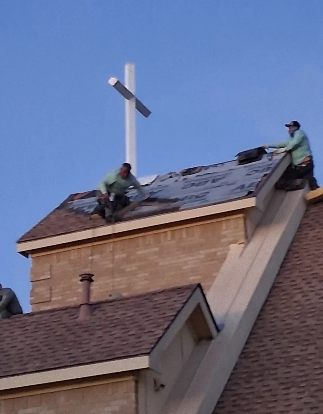 Two workers on a sloped roof repairing or installing shingles on a house, with a cross on top of a chimney in the background against a clear blue sky.