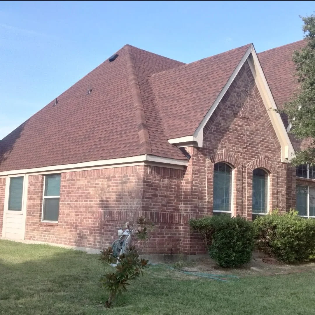 A house with a red brick exterior, multiple windows, and a sloped roof with brown shingles outdoors under a clear blue sky.