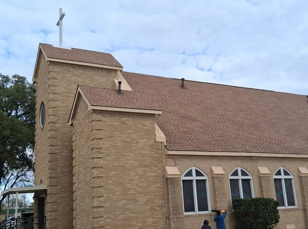 A brick church building with a steeple topped by a cross, three arched windows on the side, and a small bush in front. A person is looking through a telescope near the bushes.