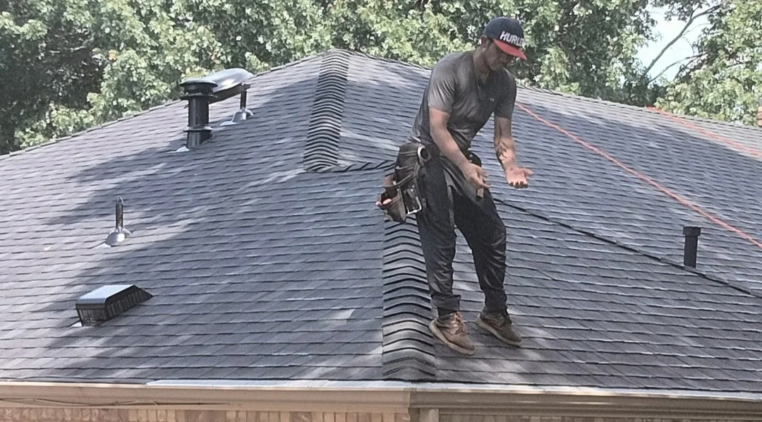 A man standing on a house roof installing or inspecting shingles, wearing a cap, gray t-shirt, black pants, and work boots, with tools attached to his belt.