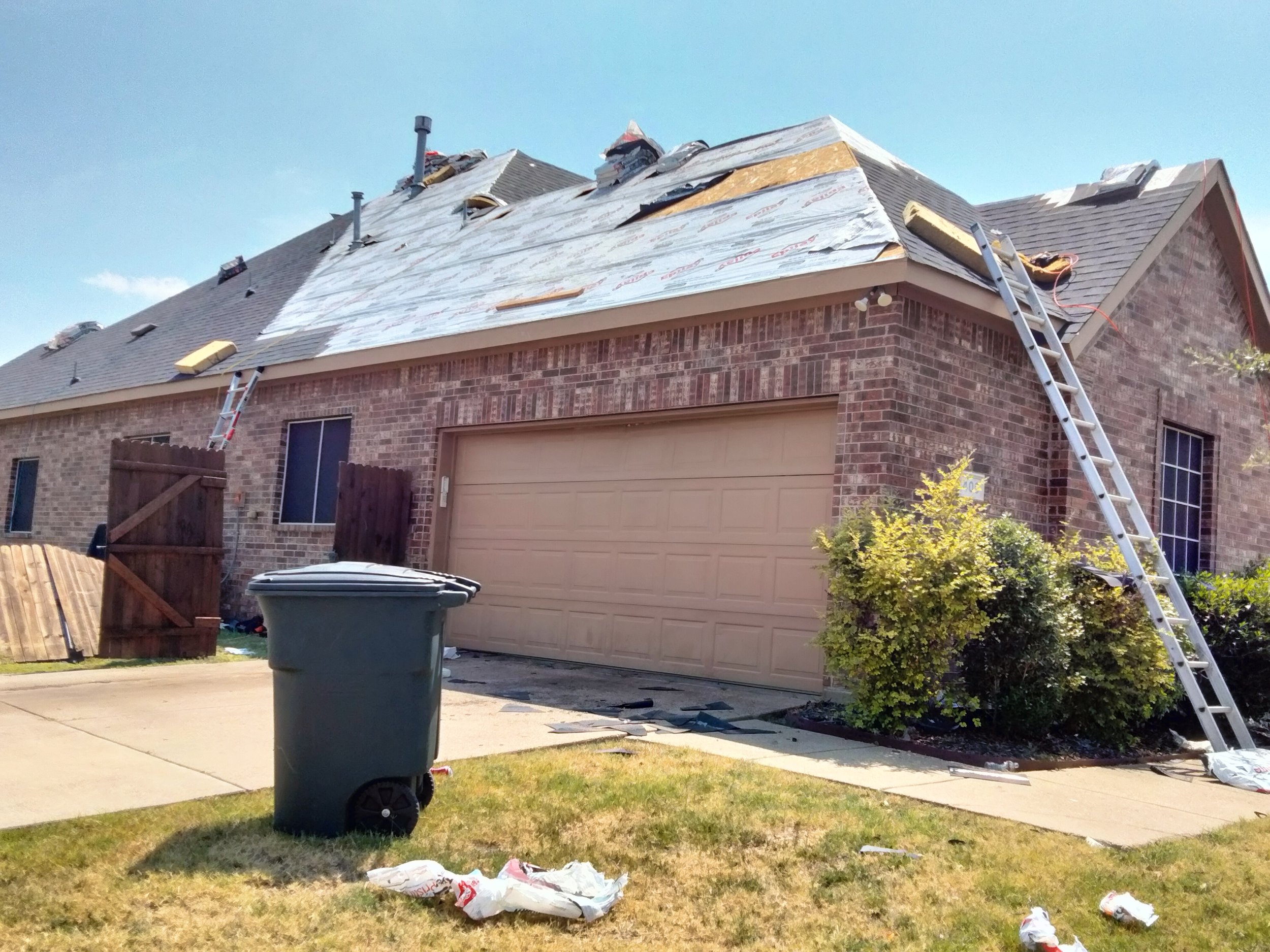 A house with its roof under repair, with ladders on both sides, some roofing materials visible, and debris scattered on the driveway and lawn under a clear blue sky.
