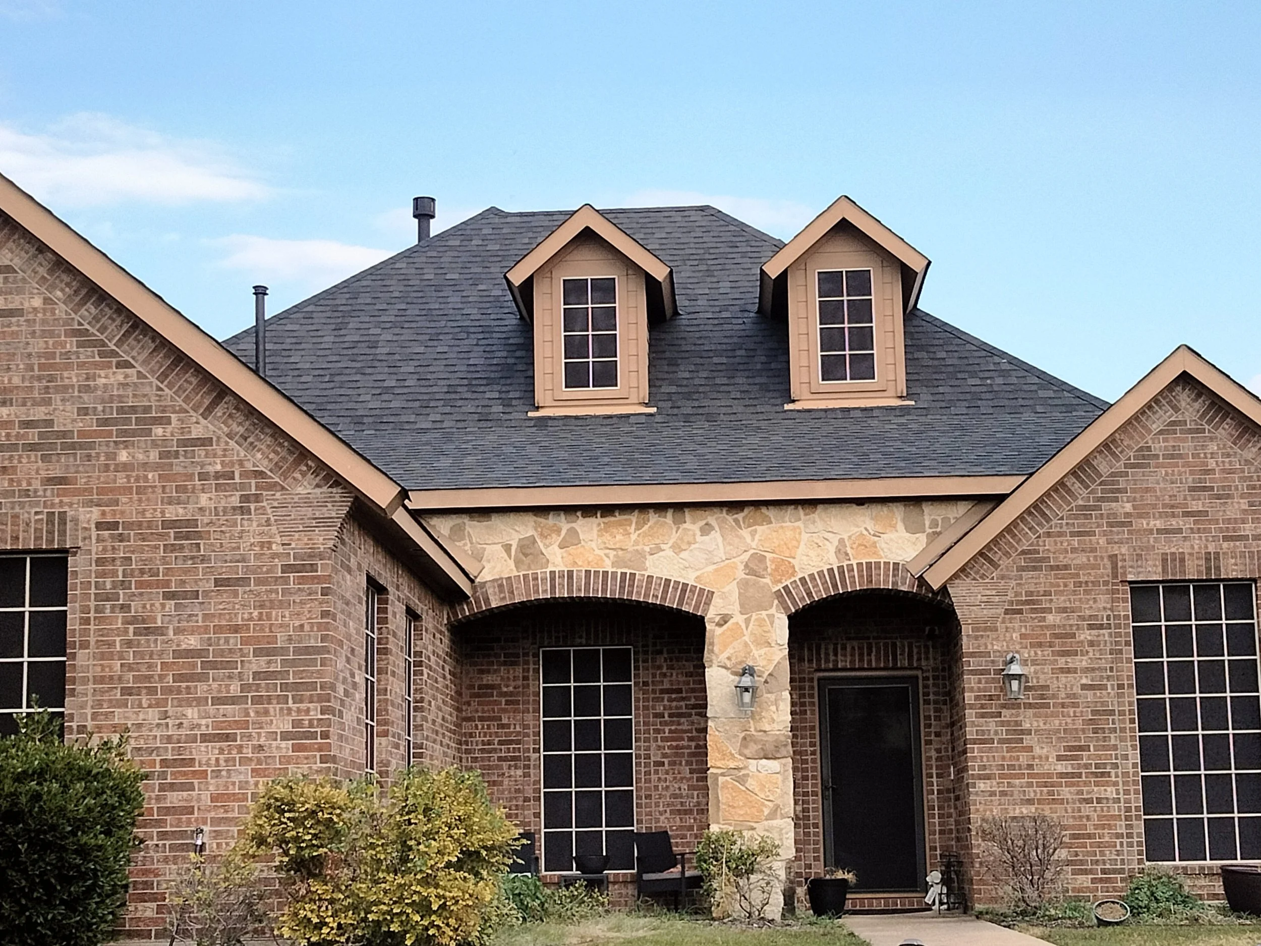 Front view of a brick house with a slate roof, two small dormer windows, a stone arch over the entrance, and a small garden with bushes and potted plants.