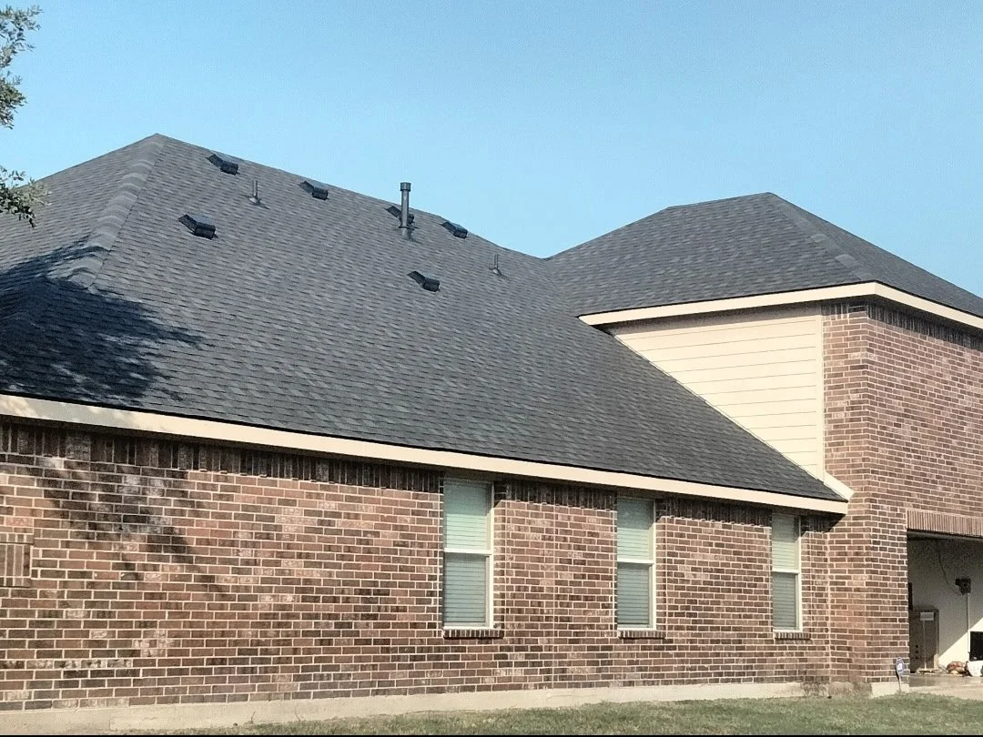 Exterior of a brick house with a dark gray shingled roof, three small windows, and a section with white siding.