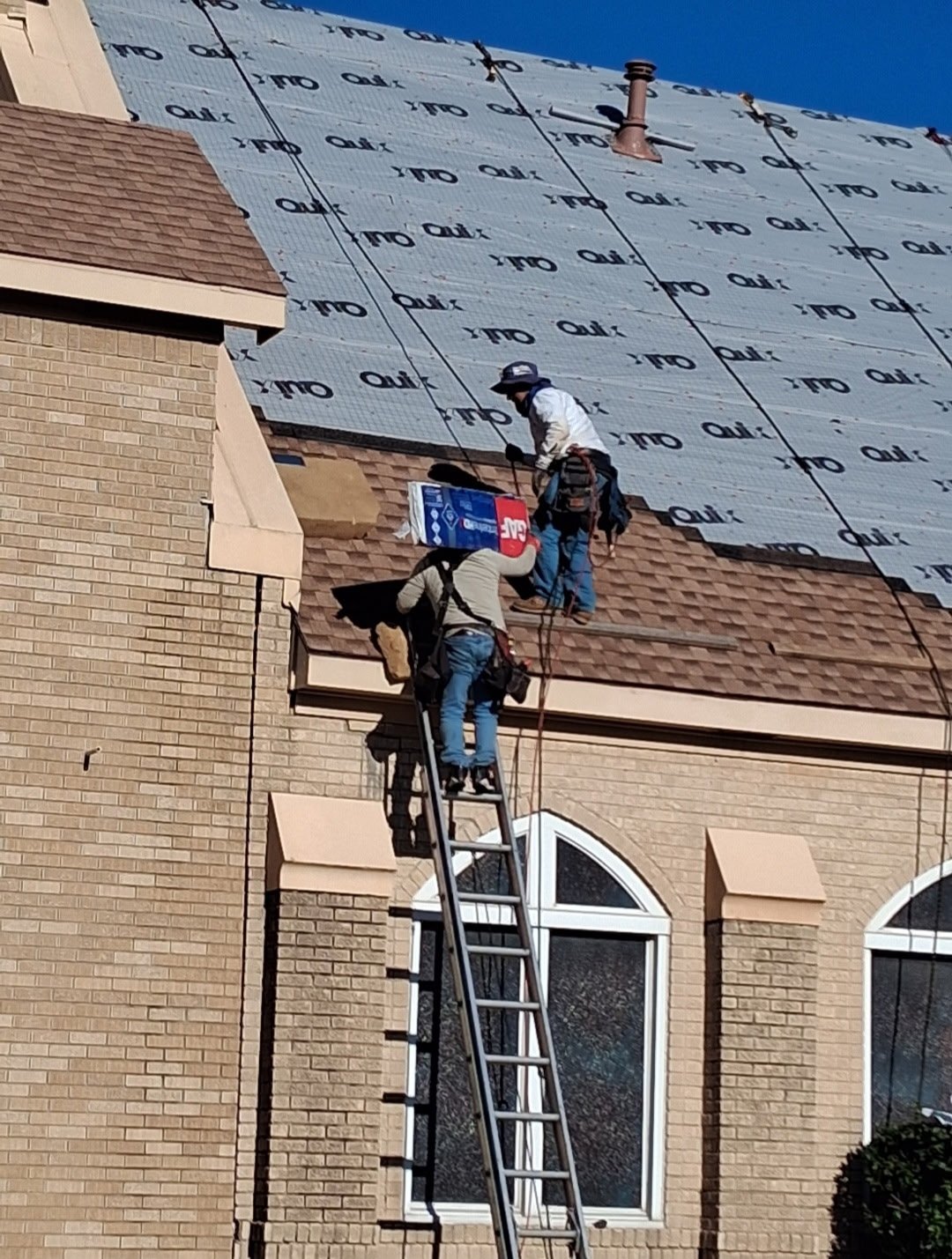 Two workers installing roofing on a house with a ladder. One worker handing a box to the other.