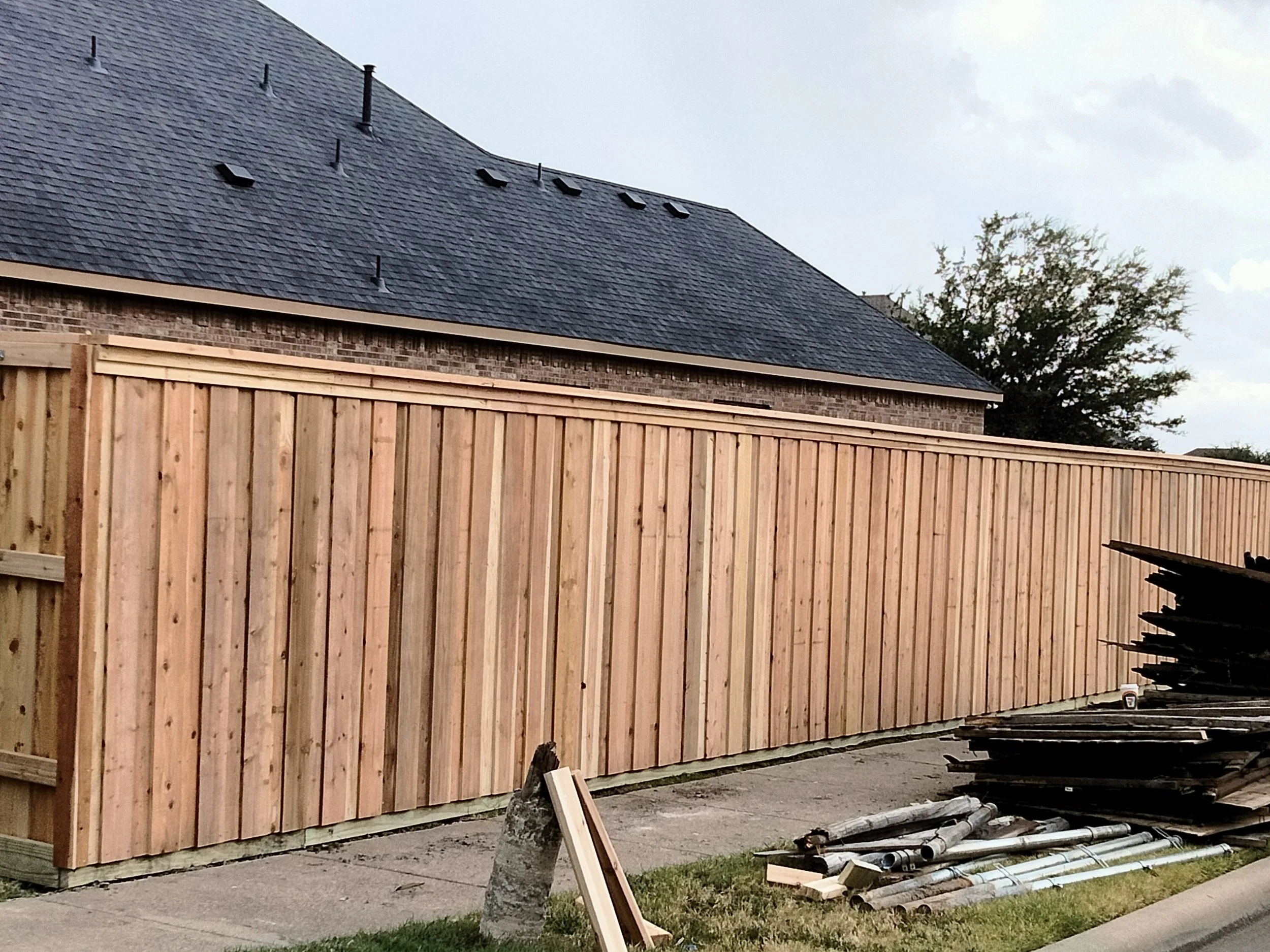 Newly constructed wooden fence along sidewalk in front of house with brick exterior and dark shingle roof. Building materials and tools are stacked on the ground beside the fence.