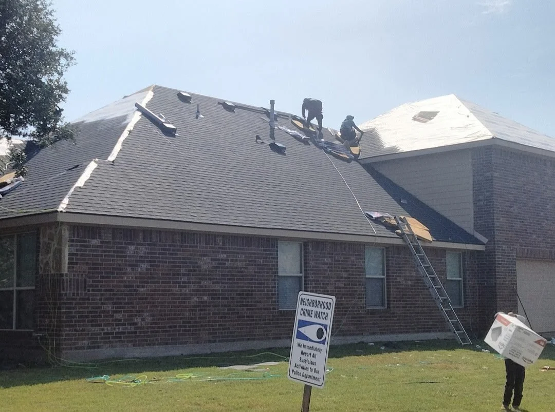 Two workers on a house roof with black roofing shingles, one kneeling and one standing. A ladder leans against the roof. Another person on the ground holds a large cardboard box, and a neighborhoood crime watch sign is visible in the yard.