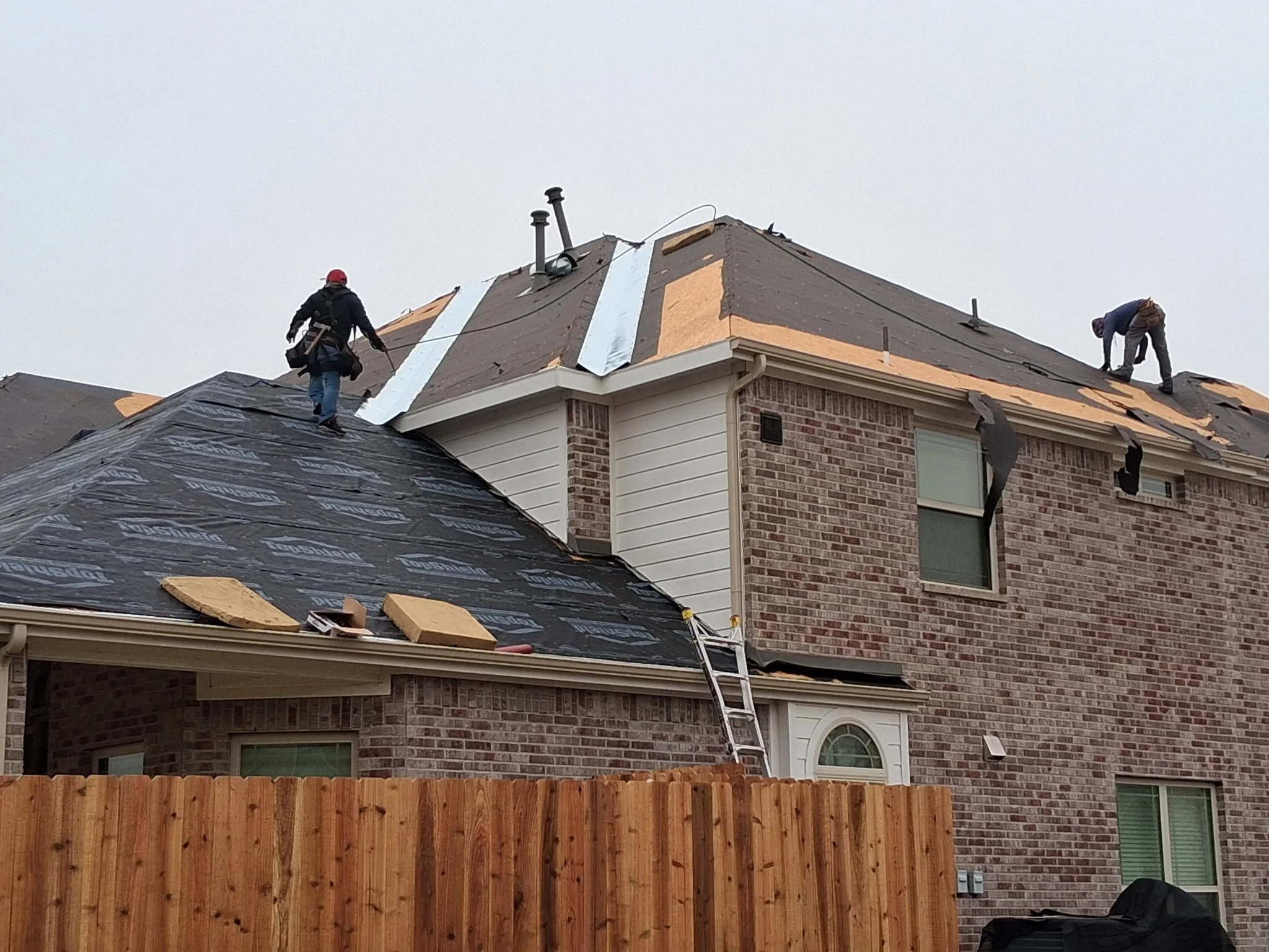 Two workers installing roofing materials on a multi-story brick house, with one worker on the left walking on the roof and another on the right crouching. There is a ladder leaning against the house and a wooden fence in the foreground.
