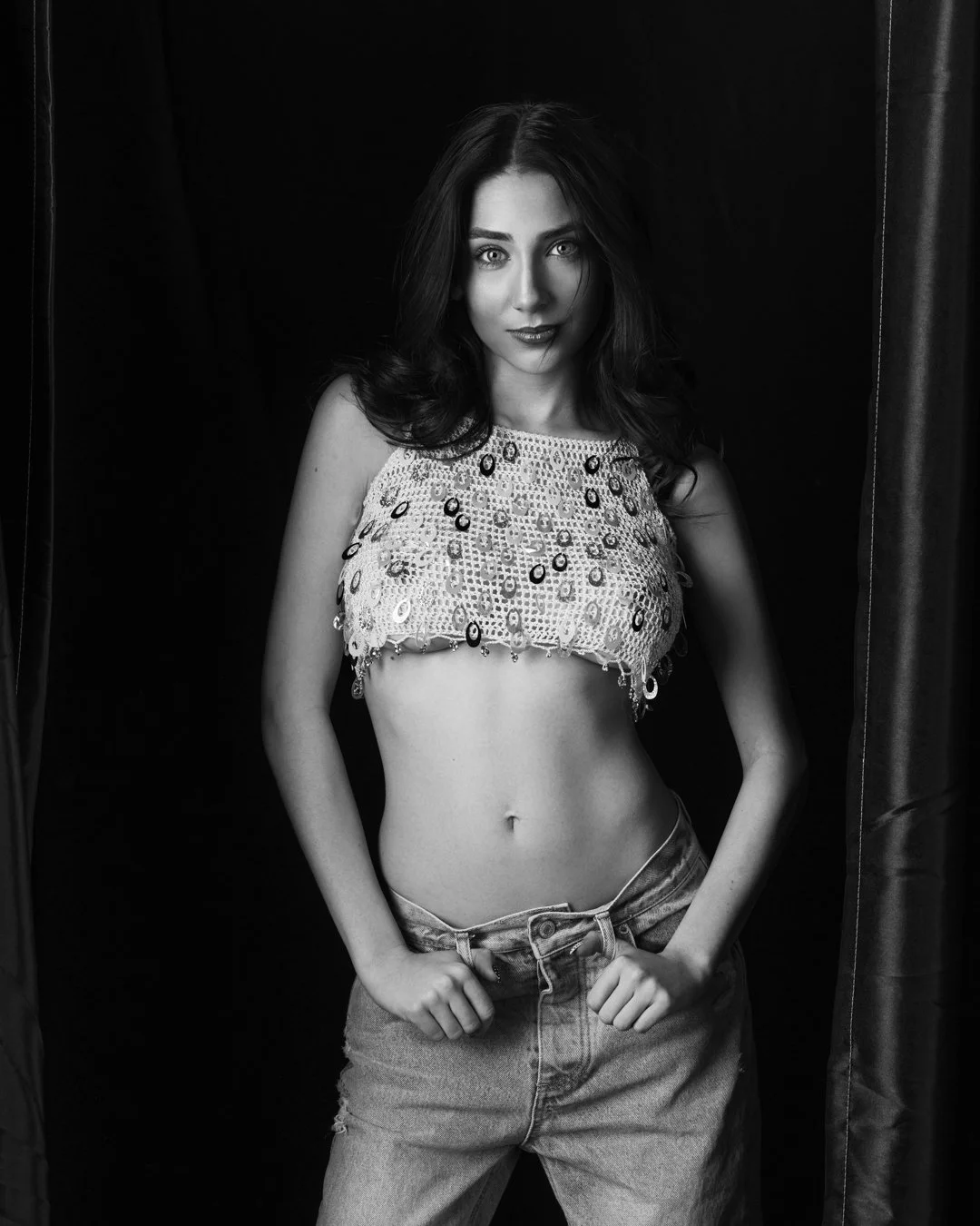 Black and white photo of a young woman with long dark hair, wearing a sequin crop top and jeans, standing in front of a dark background with curtains on either side.
