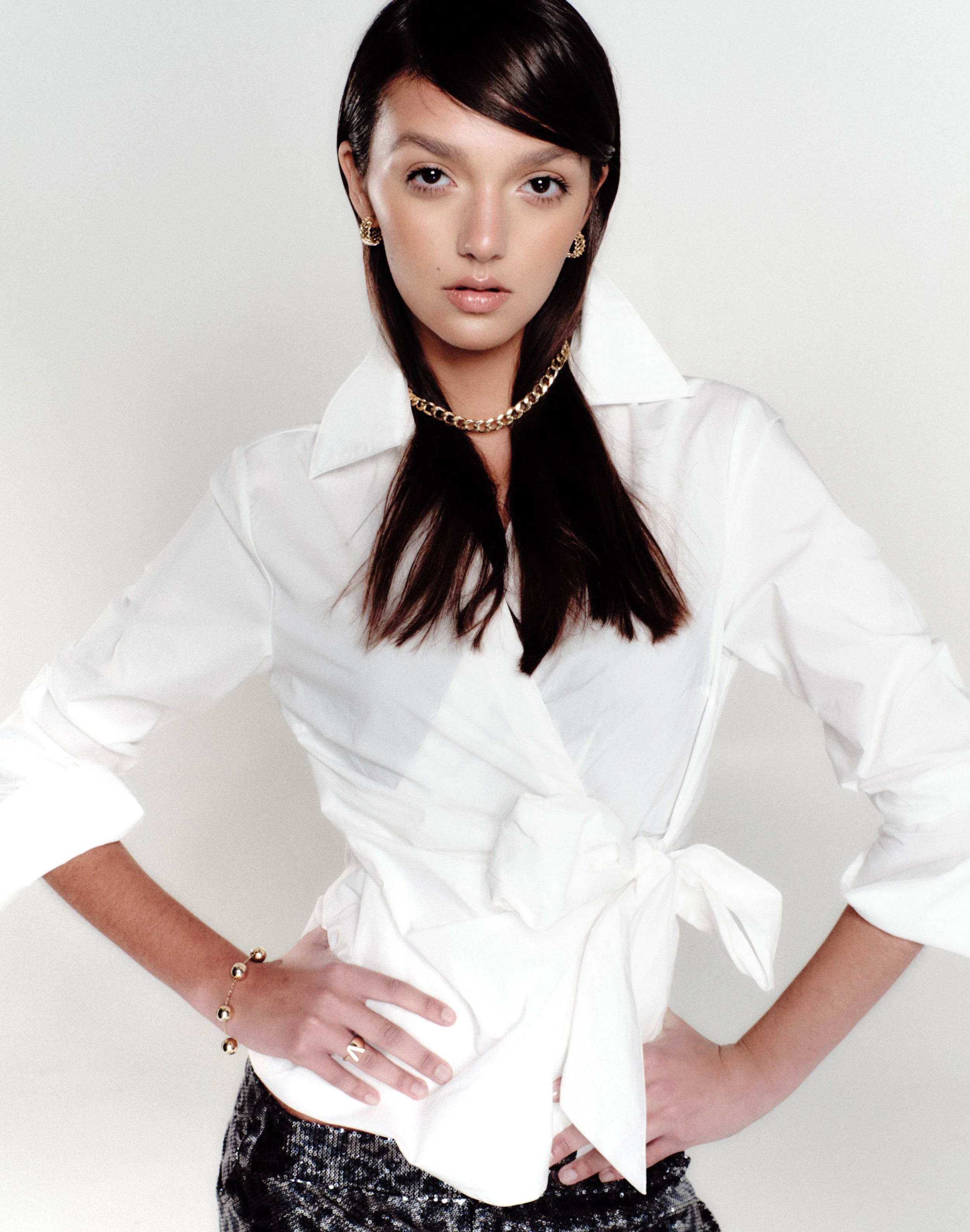 A woman with dark hair wearing a white shirt tied at the waist, accessorized with gold jewelry, standing against a plain background.