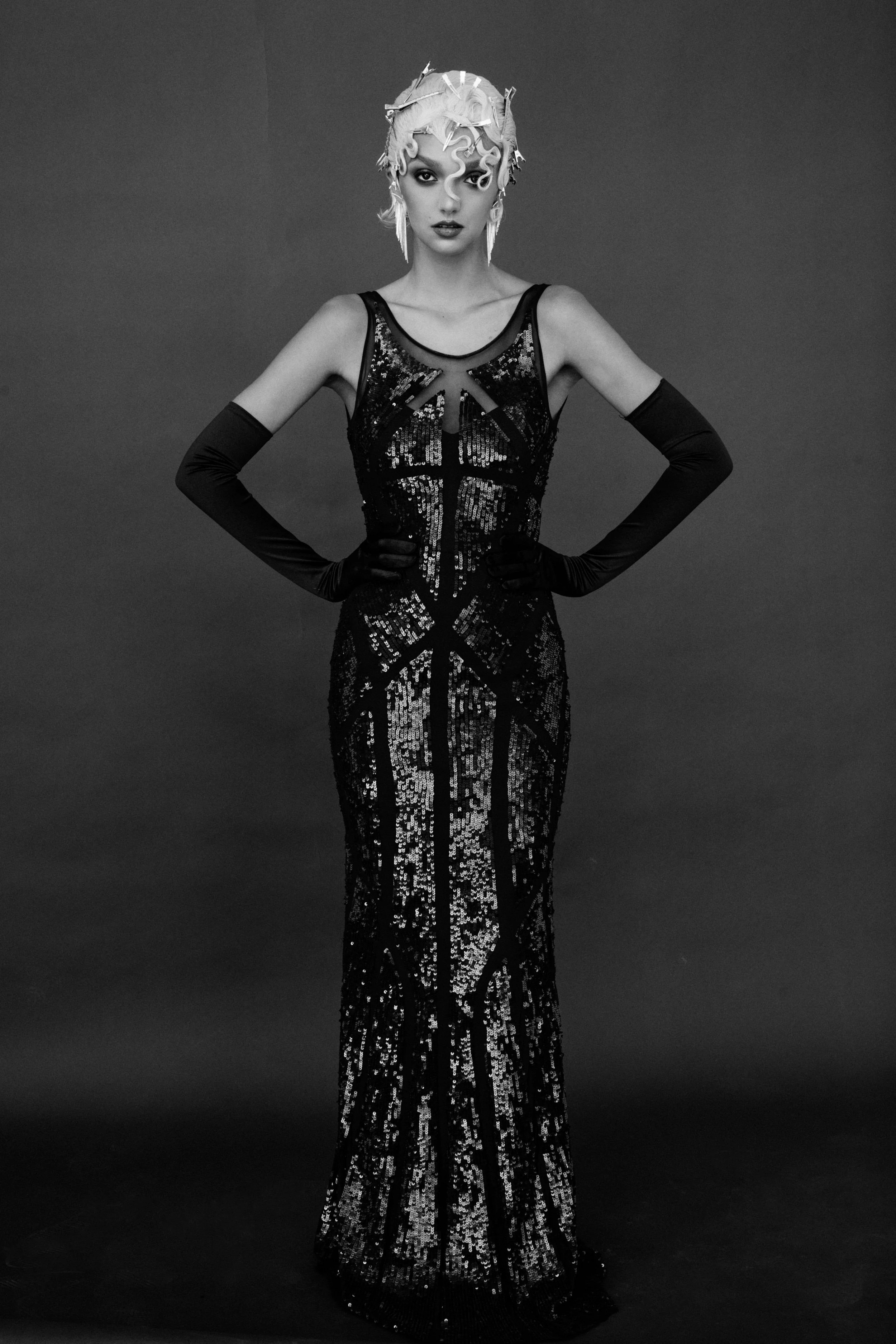 A woman in a black sequined gown with a sheer neckline, wearing long black gloves, and with hair styled in intricate curls, posing against a plain dark background.
