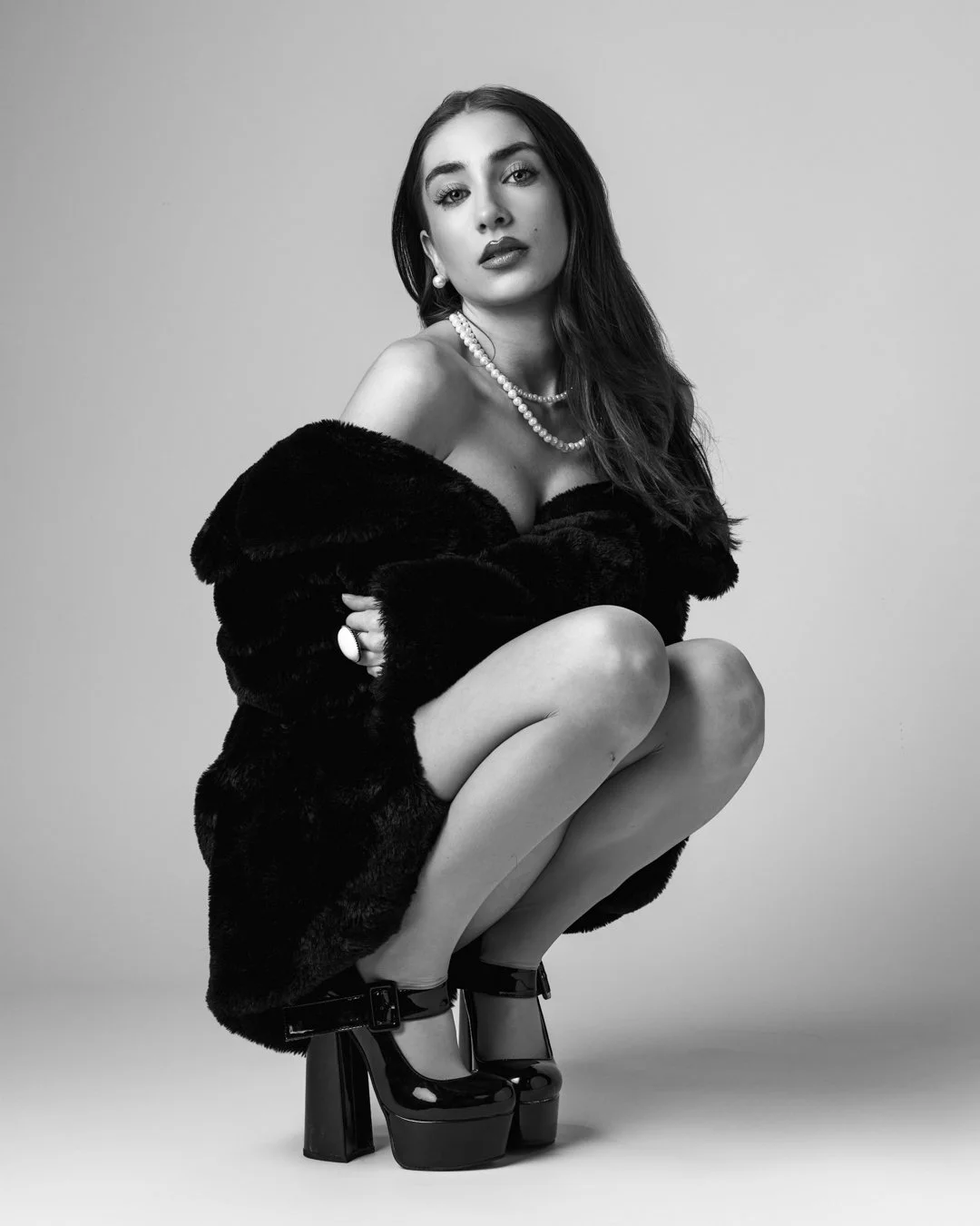 Black and white portrait of a woman with long hair, wearing jewelry, a fur coat, and high heels, squatting against a plain background.