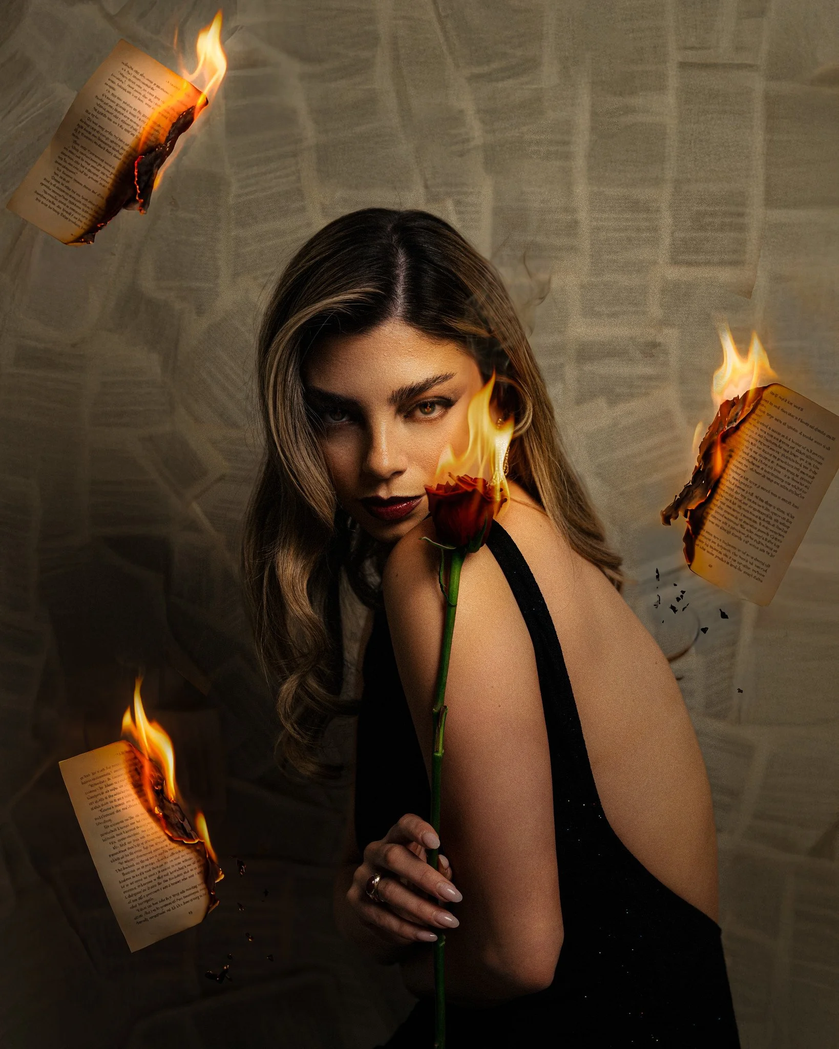 A woman with wavy brown hair holds a red rose with flames on its petals. Three open books are on fire and floating around her against a background patterned with pages.